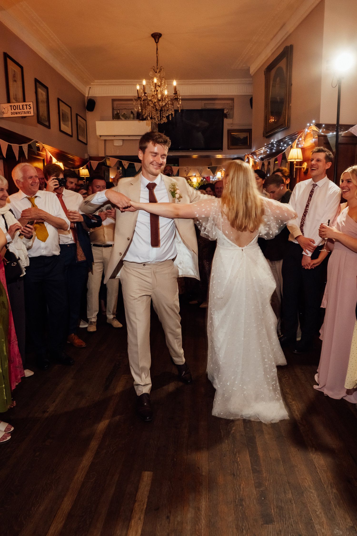 Bride in a flowing white gown dances with a smiling groom in a tan suit as wedding guests watch in a cozy pub with a chandelier.