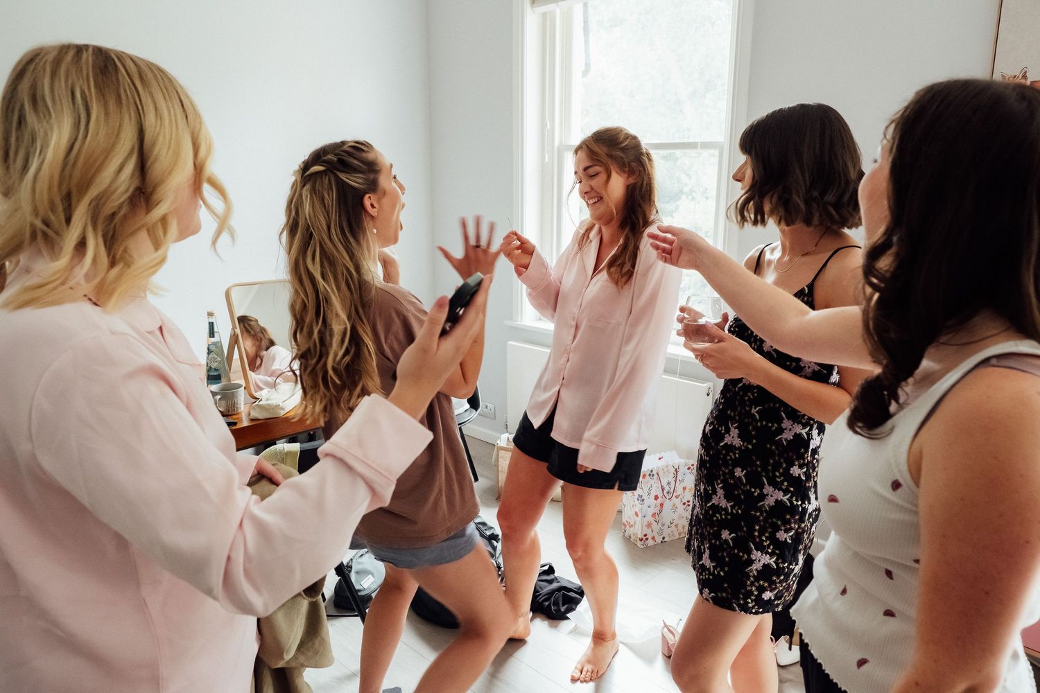 Group of women laughing and dancing together in a bright room while getting ready, wearing casual outfits and holding drinks.