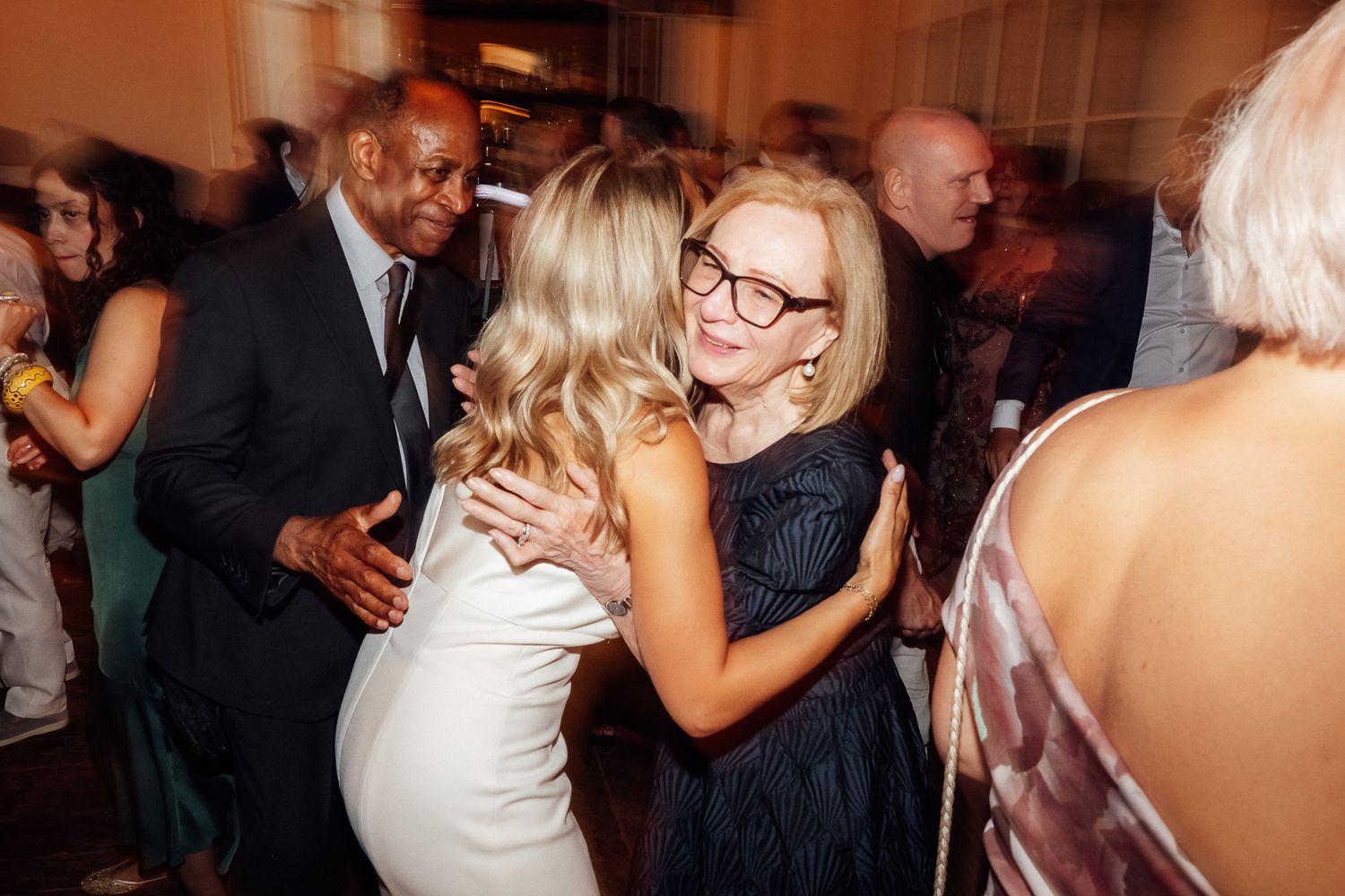 Two women in elegant dresses embrace warmly at a crowded indoor party while a smiling man in a dark suit and other guests look on.
