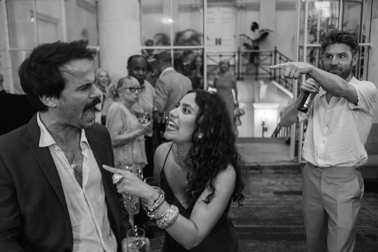 Black and white photo of a woman with curly hair pointing and laughing beside a man in a blazer while another man speaks into a microphone.