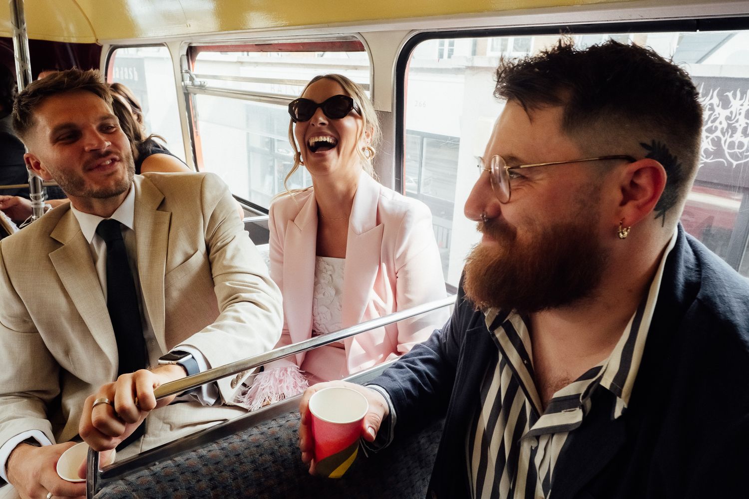Three friends laugh and celebrate inside a vintage double-decker bus, one holding a red cup, dressed in stylish wedding party attire.