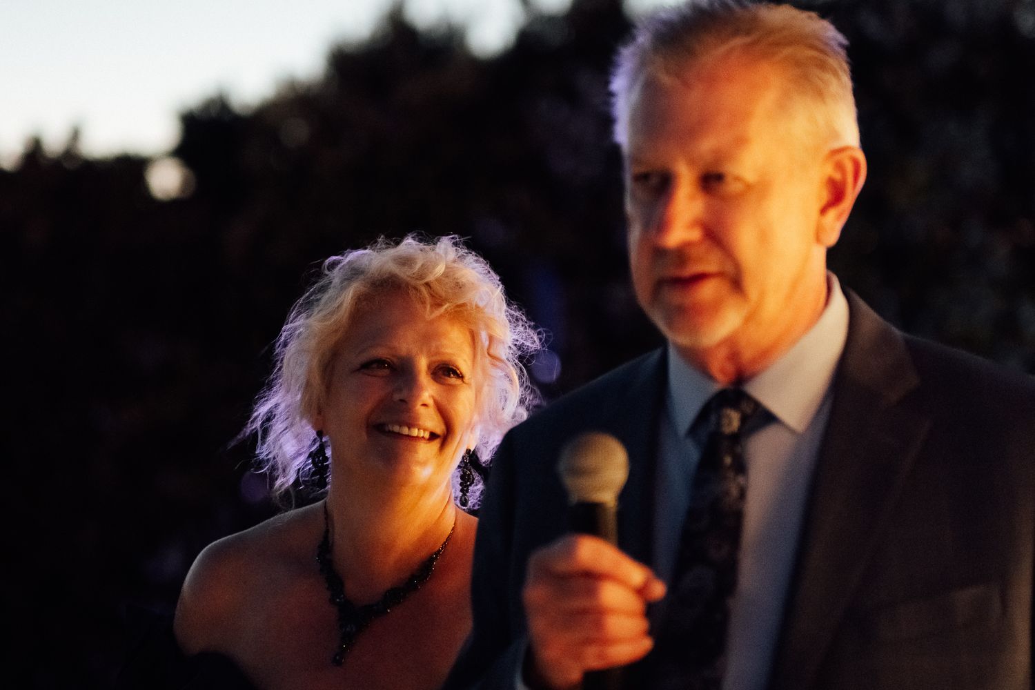 Man speaking into microphone with smiling woman beside him at an outdoor evening event.