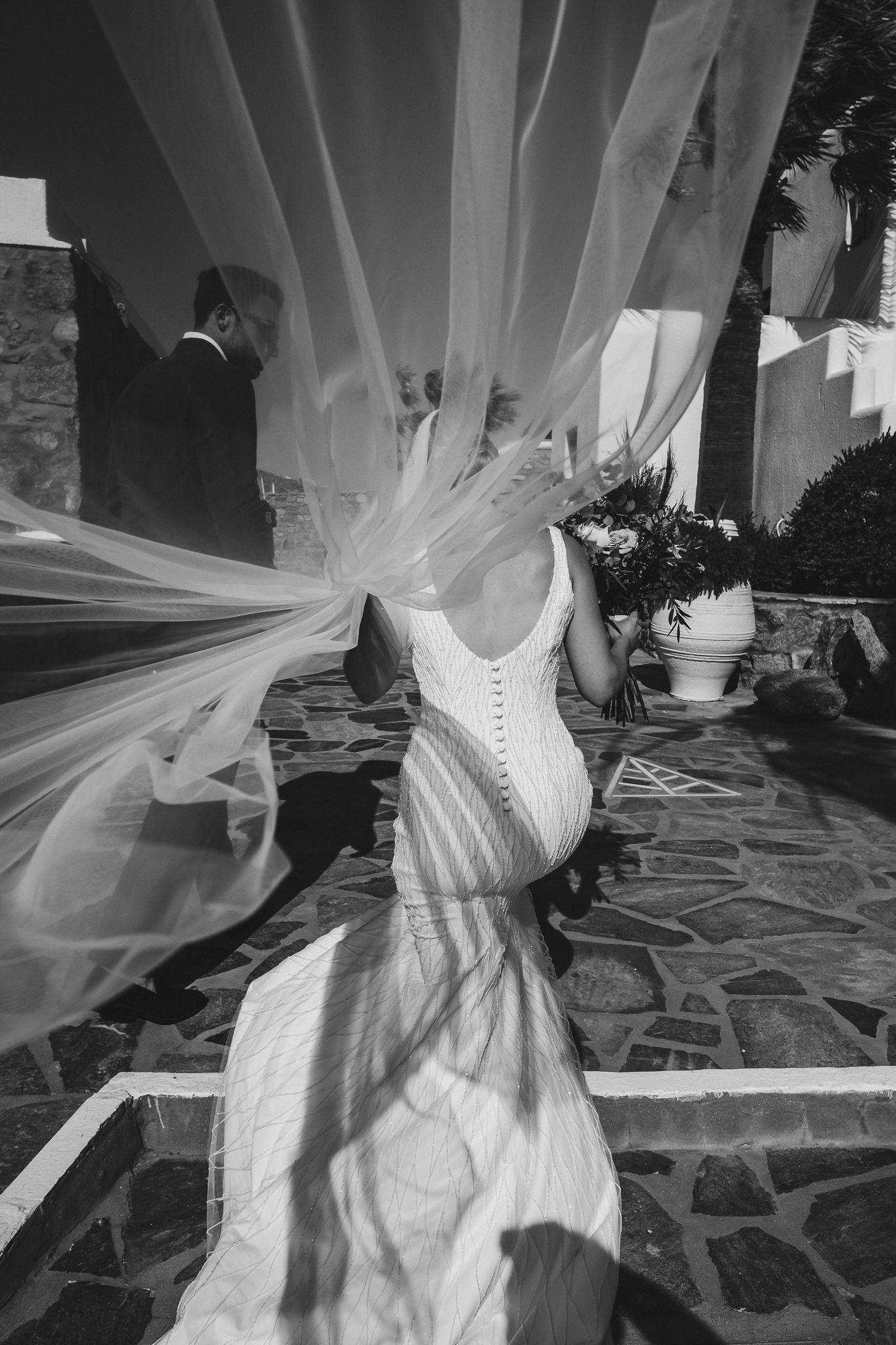 A flowing white corset wedding dress with tulle veil creates an ethereal silhouette against a stone wall backdrop.