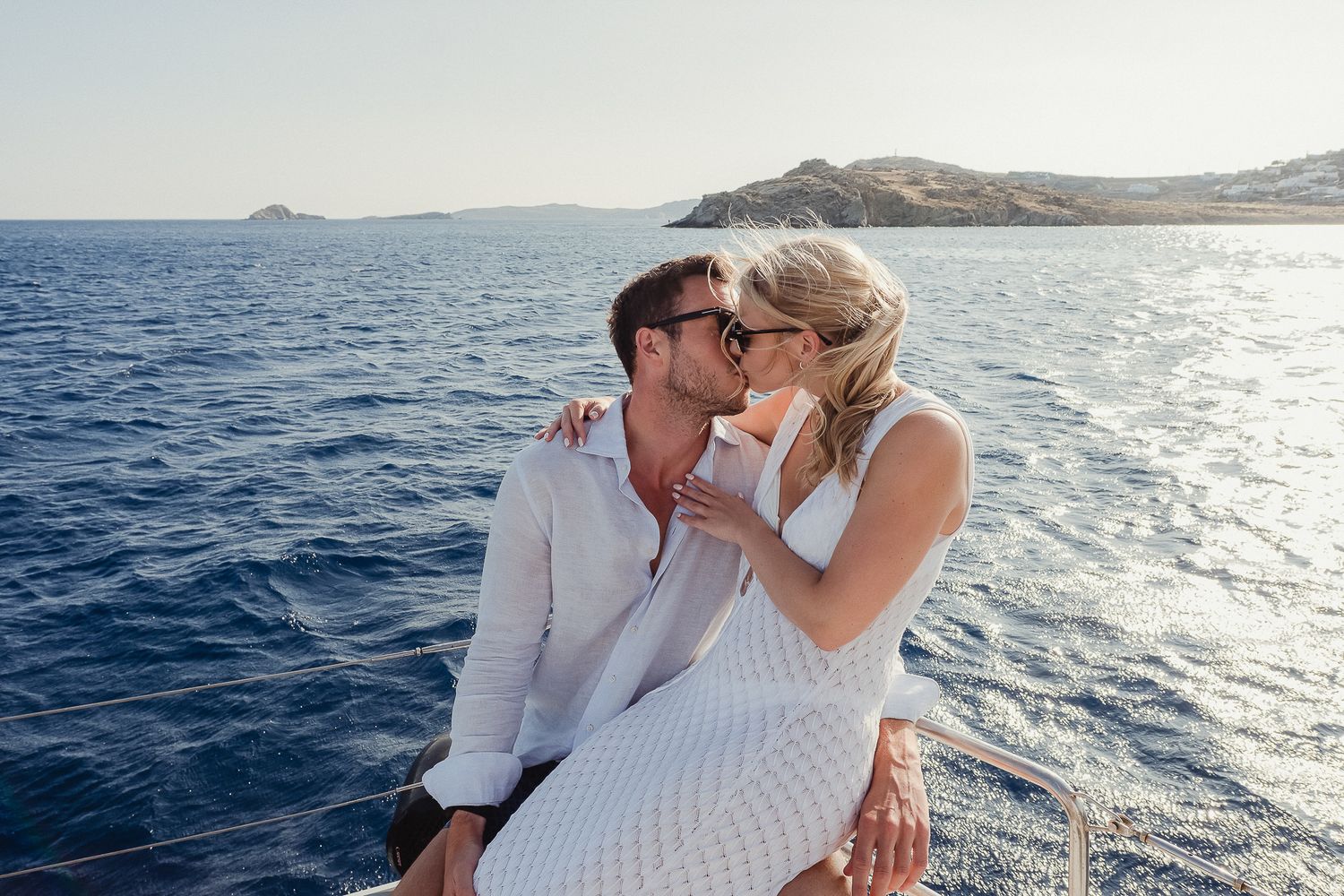 A couple shares a romantic moment together on a boat with a scenic ocean and mountain backdrop.