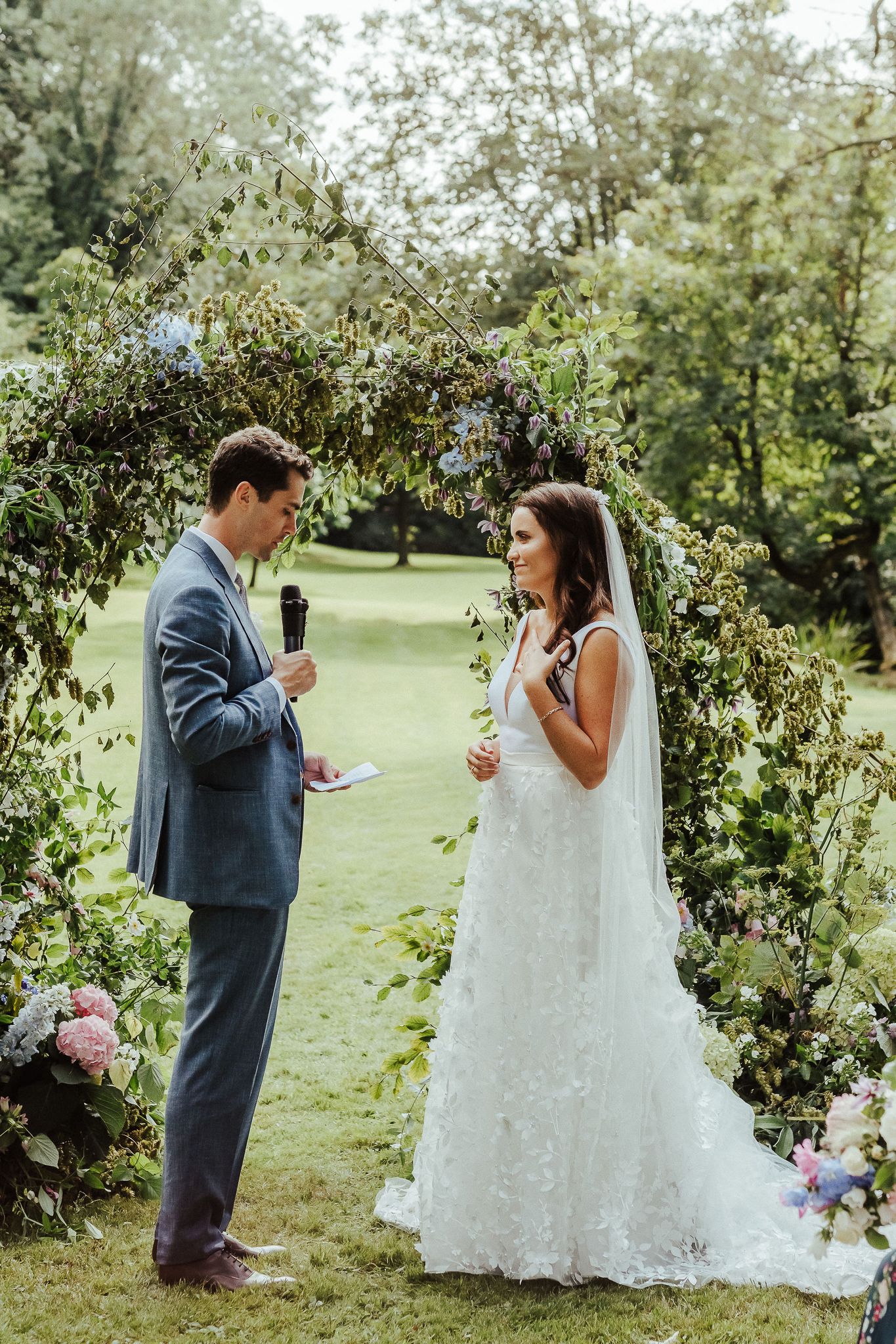 Bride and groom exchange personal vows during their outdoor garden ceremony under a floral archway.