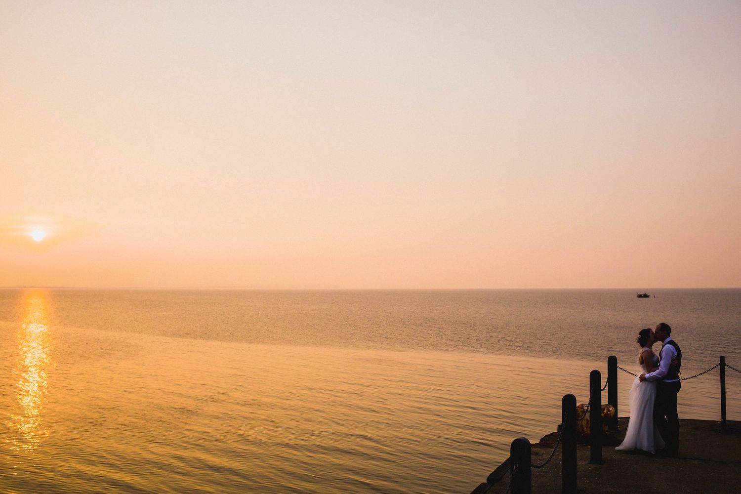 A couple stands on a dock overlooking a golden sunset across calm waters at dusk.