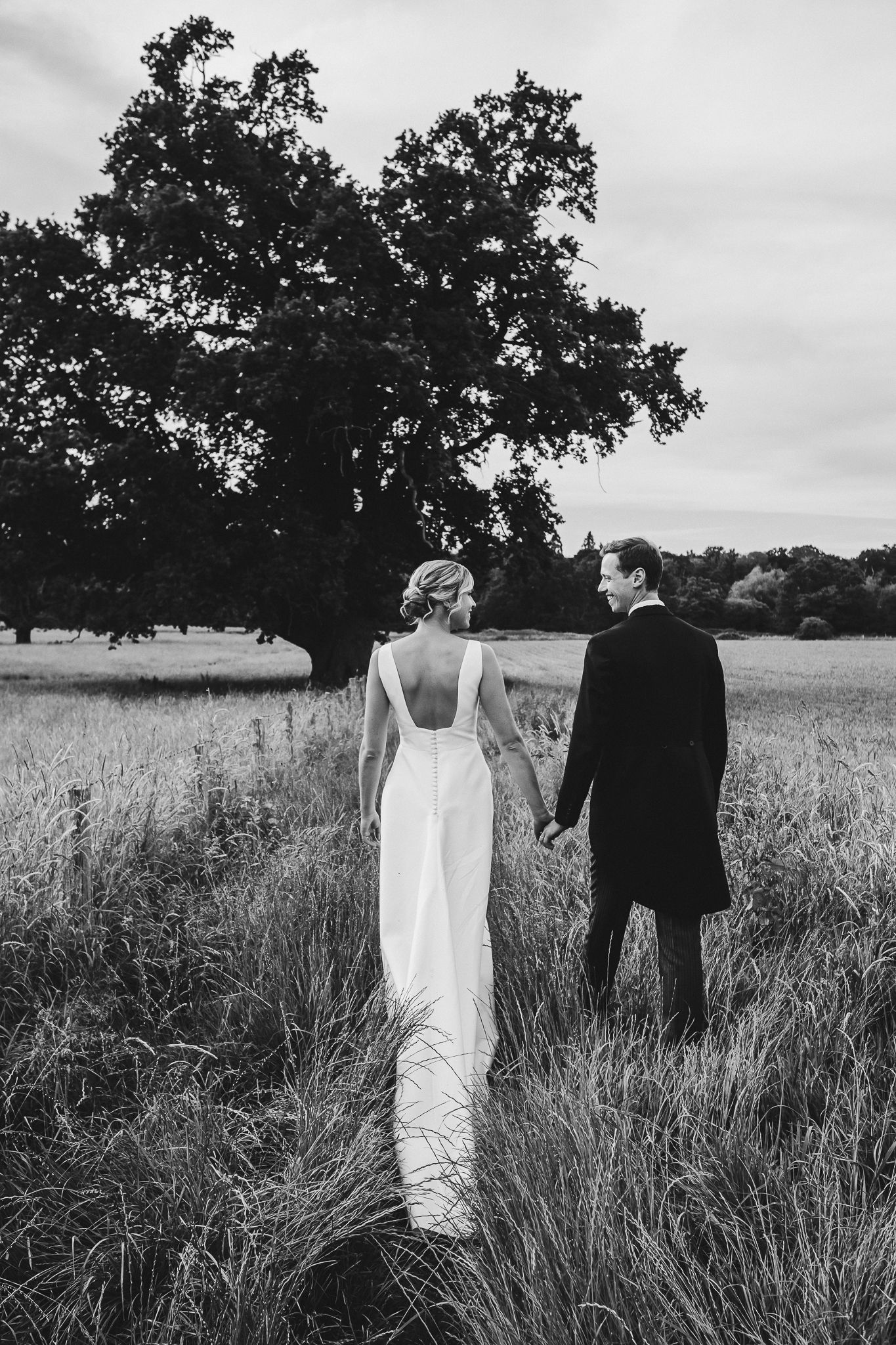 A black and white photo of newlyweds standing together in a field with a majestic oak tree.