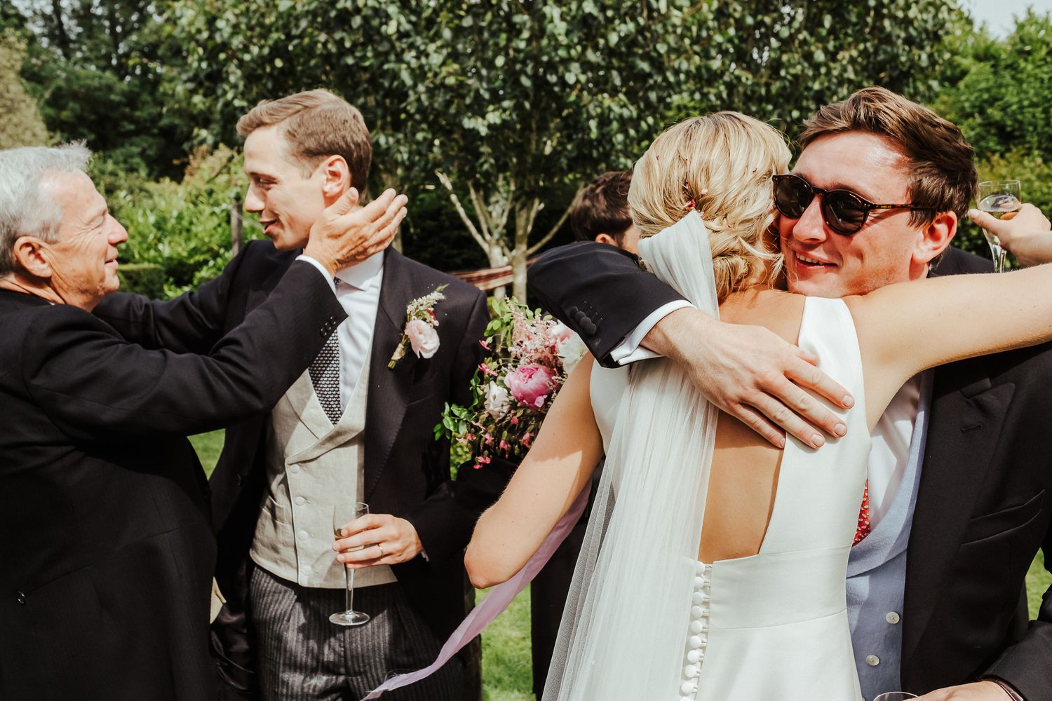Wedding guests laugh and celebrate with warm hugs and embraces in a sunny outdoor garden setting.