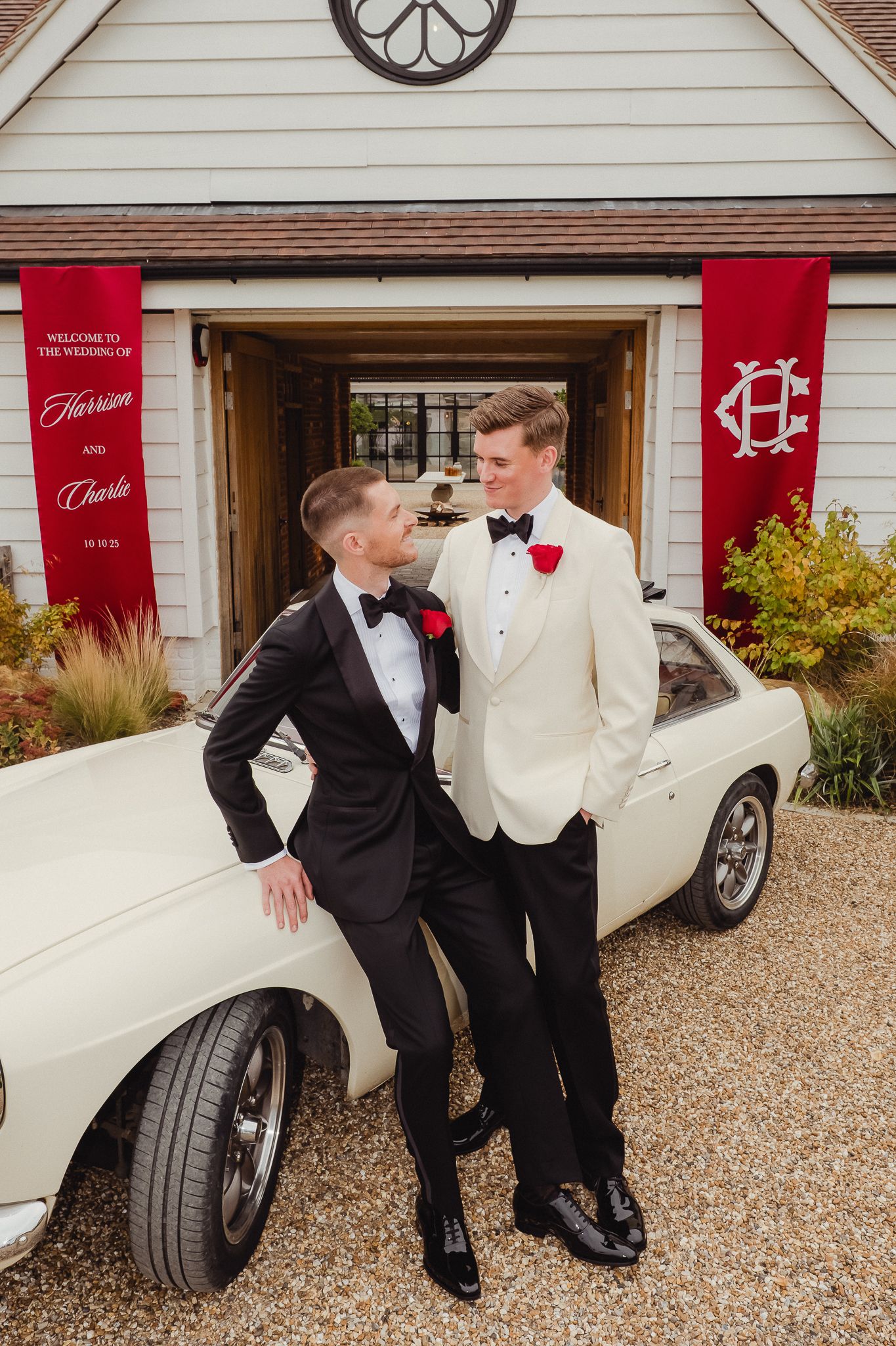 Two people in formal attire pose playfully with a white classic car outside a house with red banners.