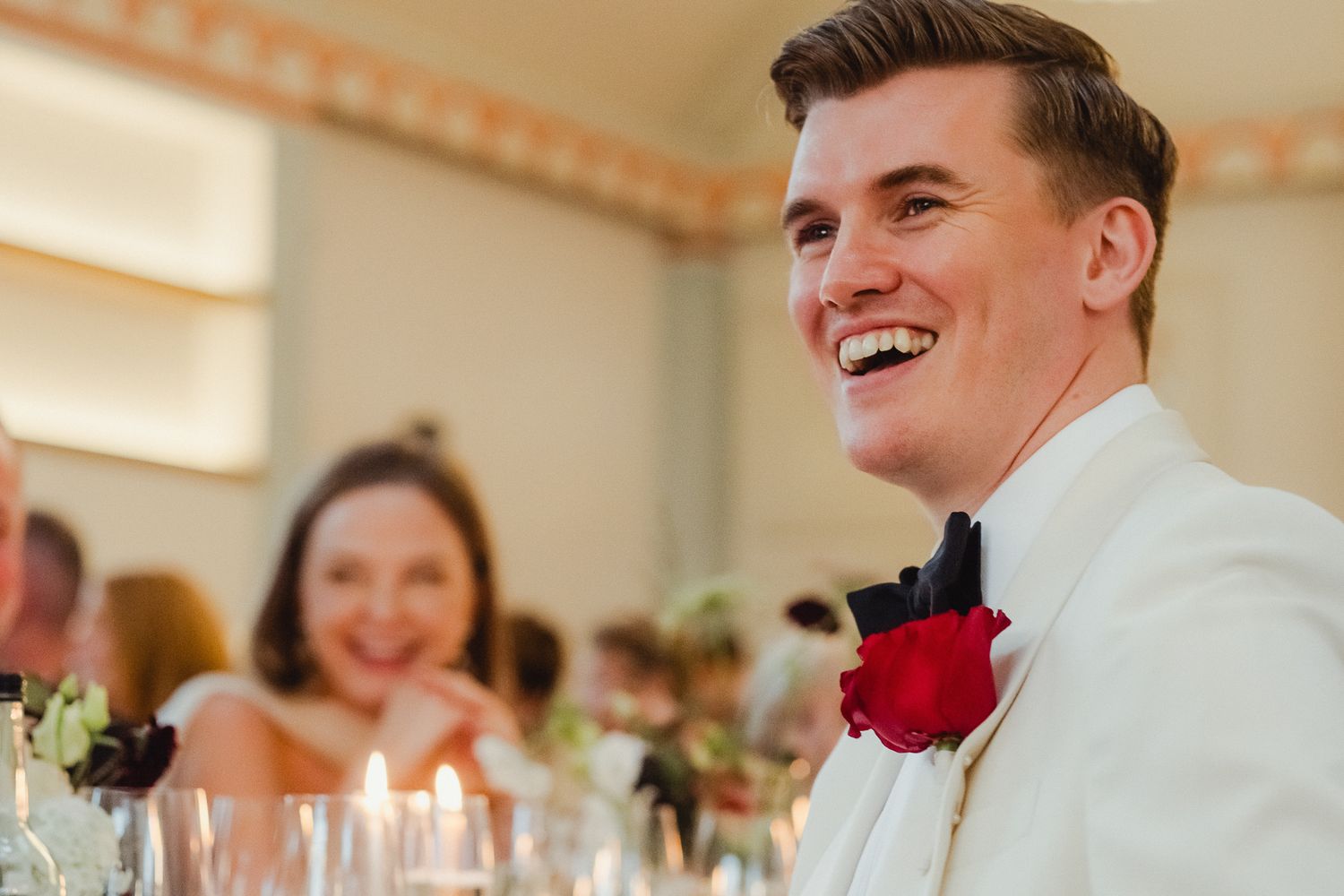 People celebrate at a wedding reception with lit candles and red roses decorating elegant white tables.