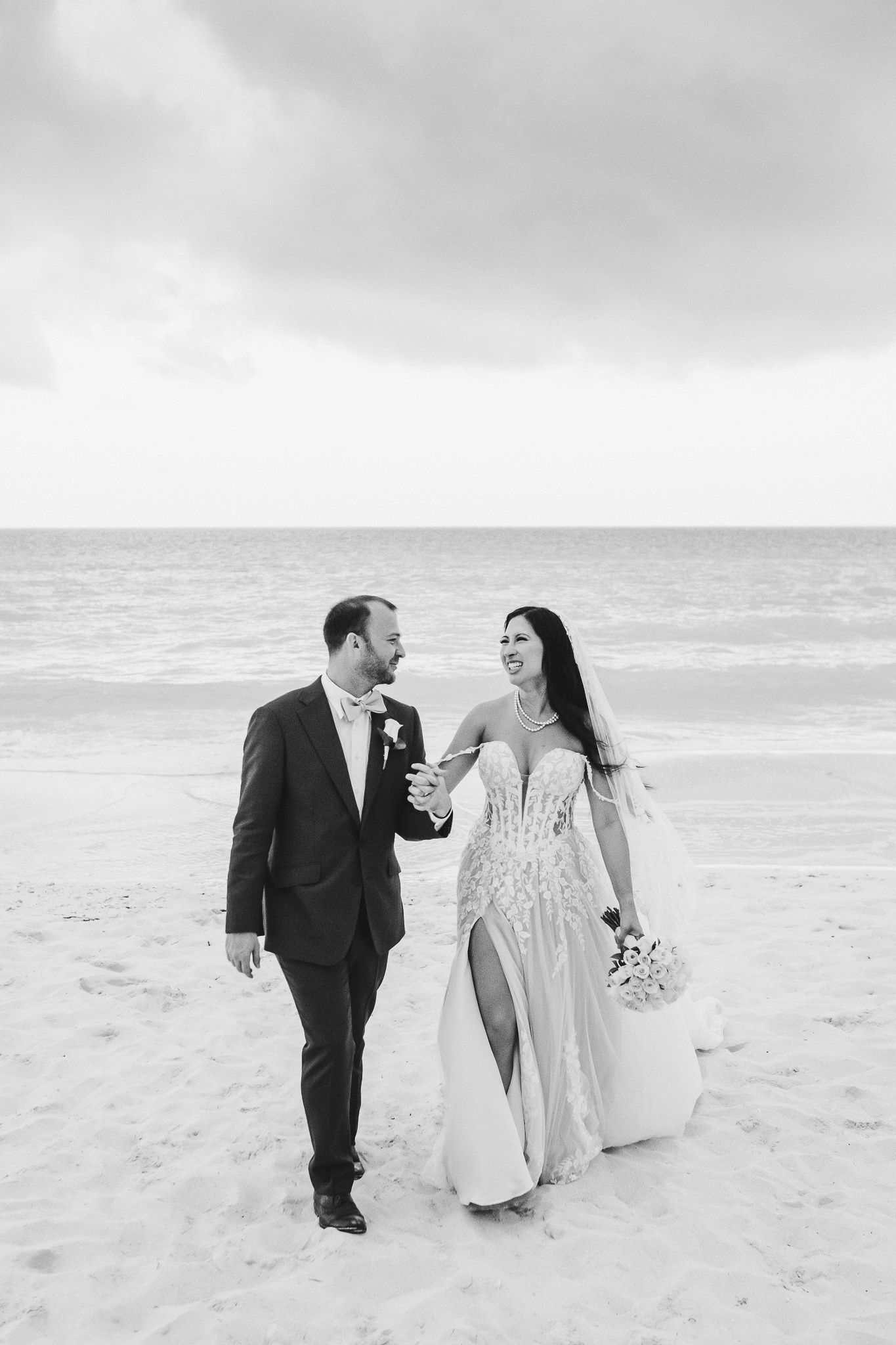 A couple in wedding attire walks together and laughs on a cloudy beach with waves in the background.