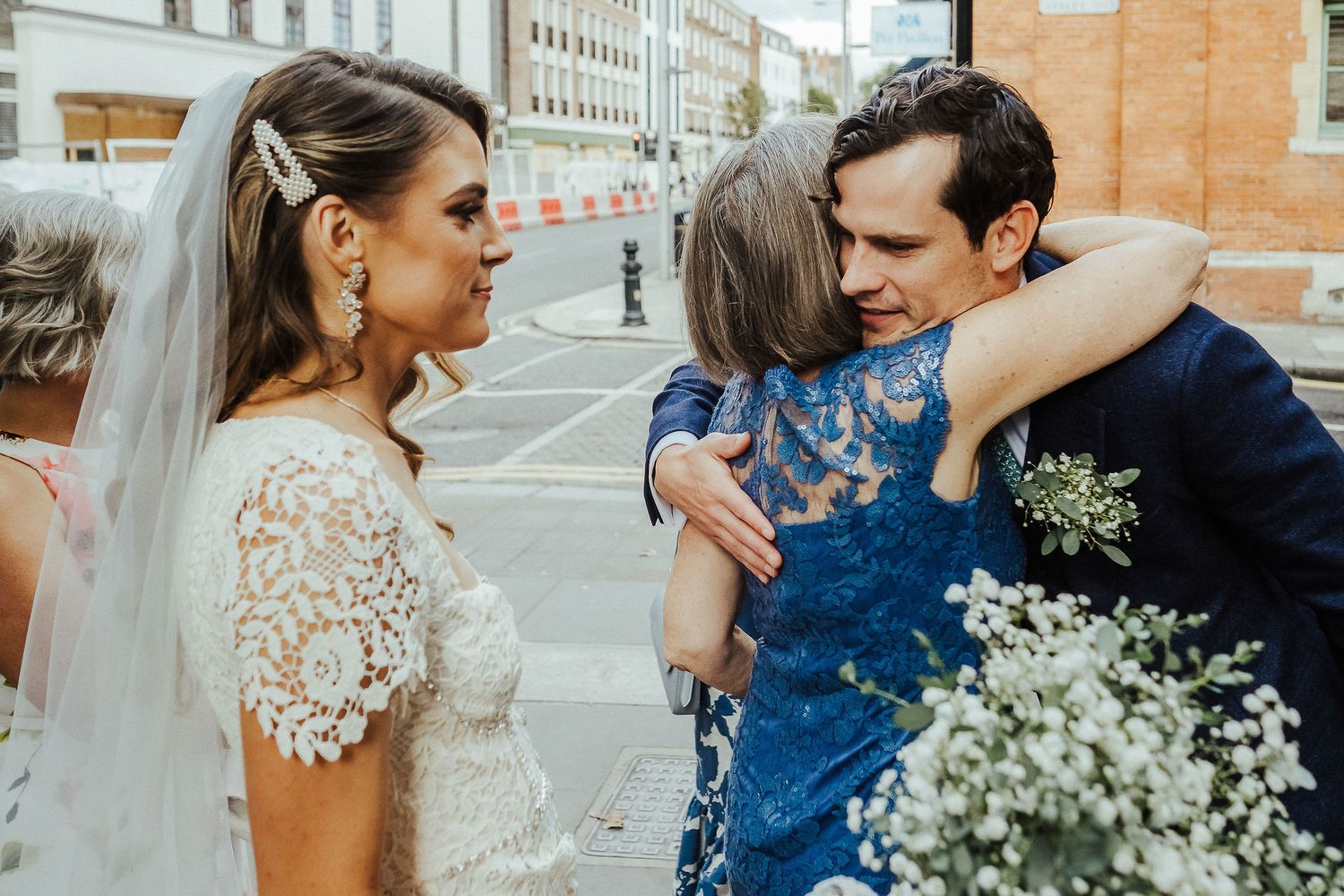 A couple in wedding attire embraces while standing on a city street, with white flowers in view.