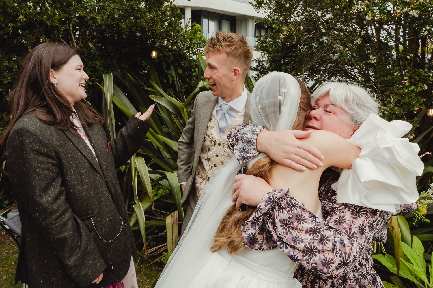 Three people share a candid laughing moment together in a garden setting during a wedding celebration.