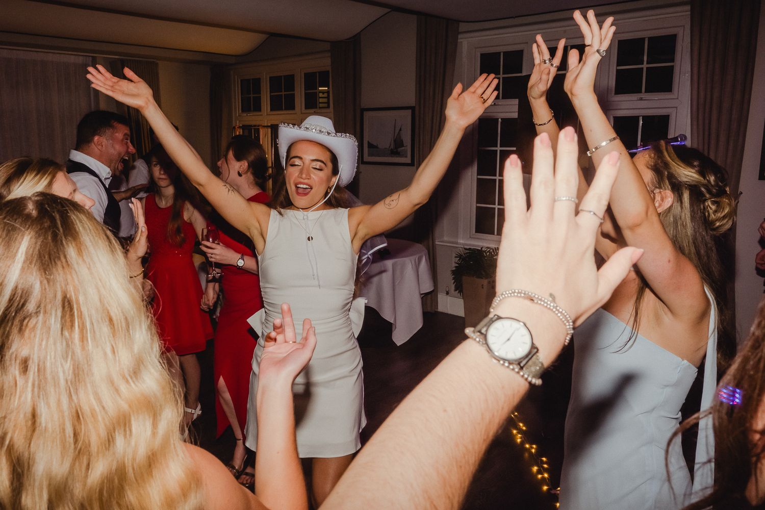 Wedding reception guests dance and celebrate with raised arms on a crowded dance floor at an indoor venue.