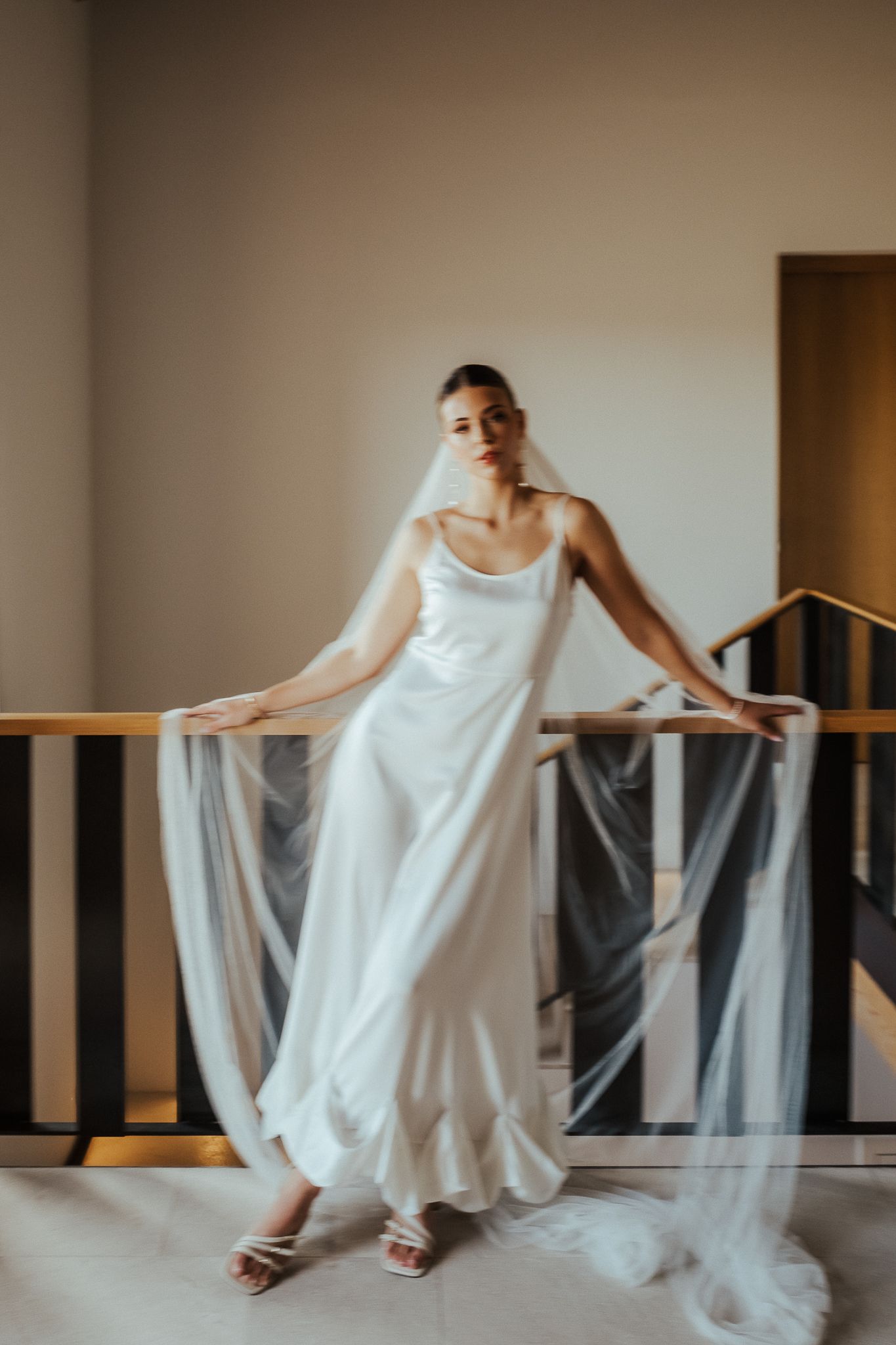 A bride in a white wedding dress holds flowing tulle fabric while posing in an elegant interior space.