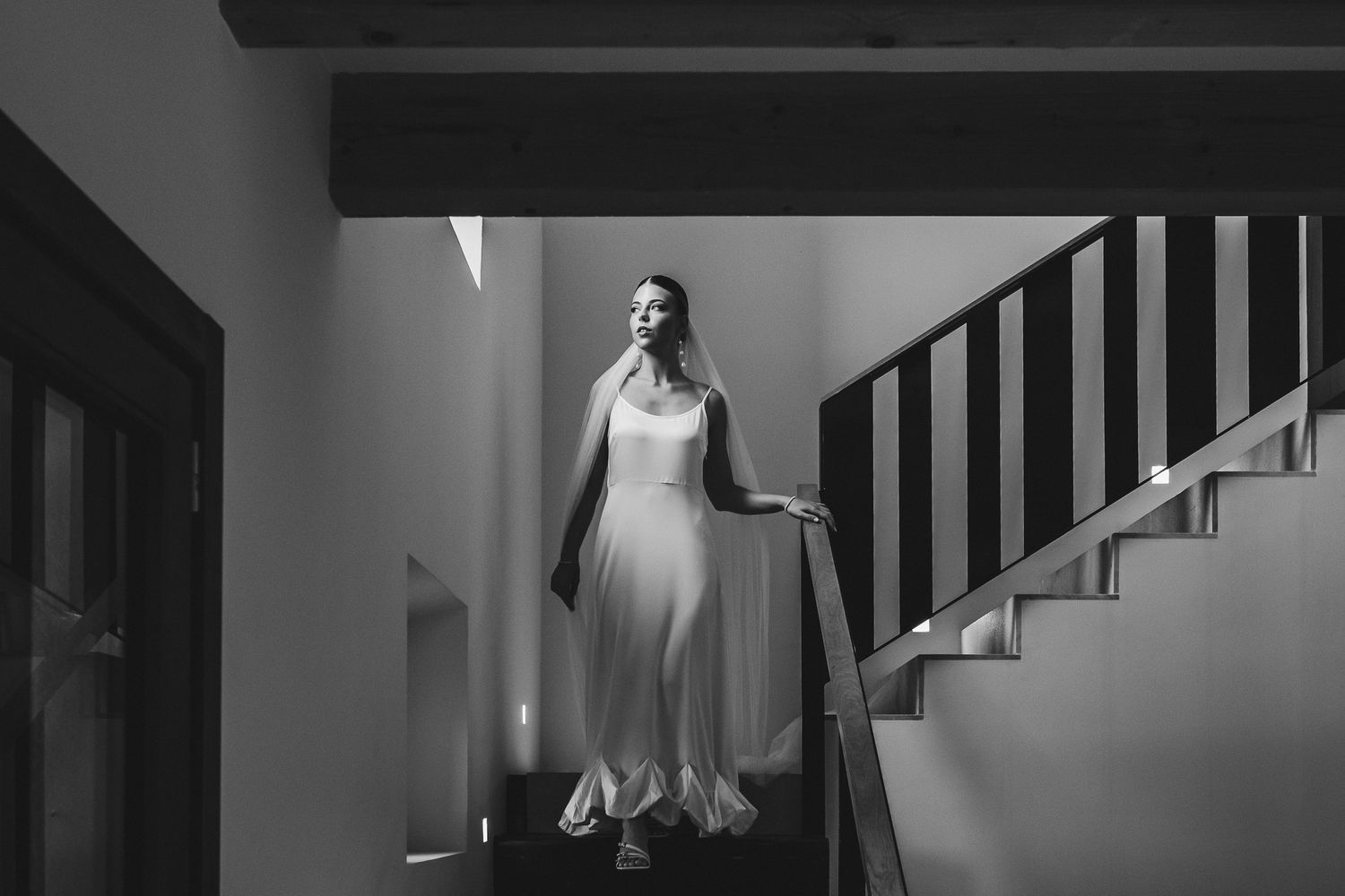 A figure in an elegant white wedding dress stands gracefully on stairs in a dramatic black and white photograph.
