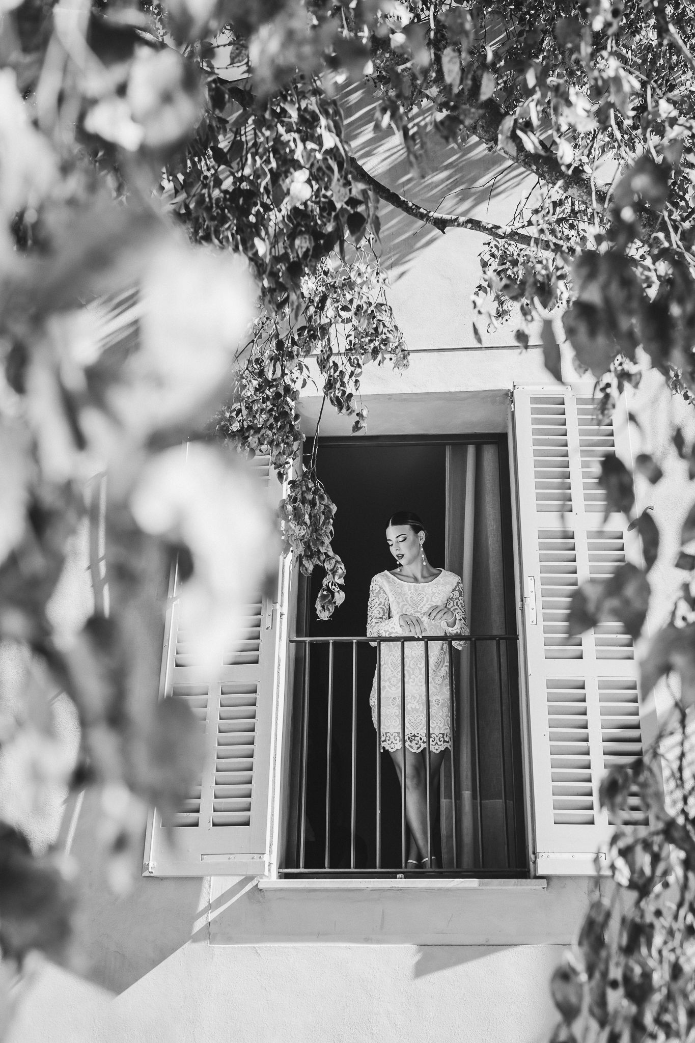 A person looks out through an open window with white shutters framed by blurred tree branches in black and white.