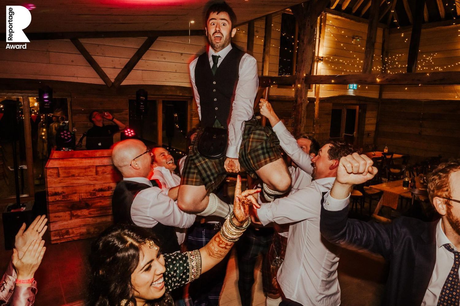 A person being lifted up by a group of party guests during a lively celebration in a rustic venue with warm lighting.