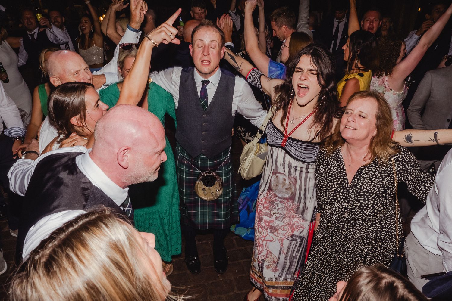 A lively crowd of people dancing and celebrating at a party with energetic expressions and raised arms.
