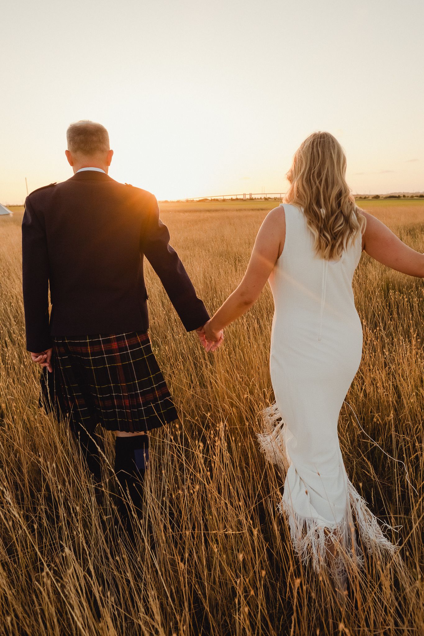 A couple walks hand in hand through a golden wheat field at sunset wearing formal attire.