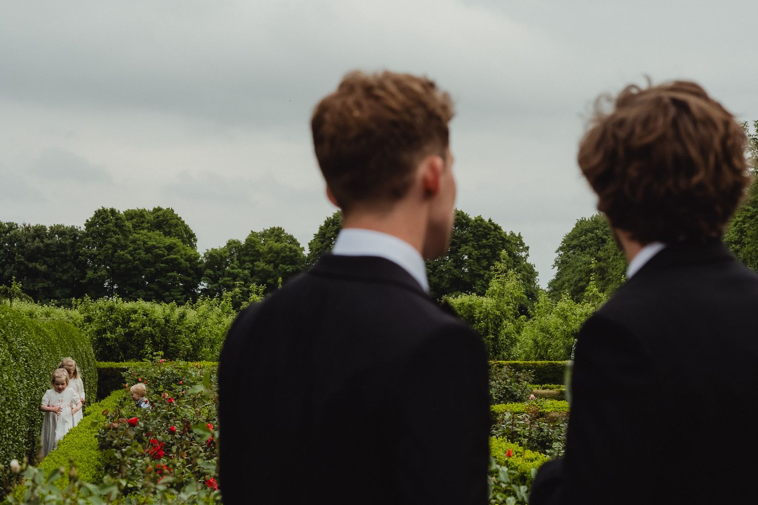 Two people in black suits view a garden wedding ceremony from a distance with trees and flowers in the background.