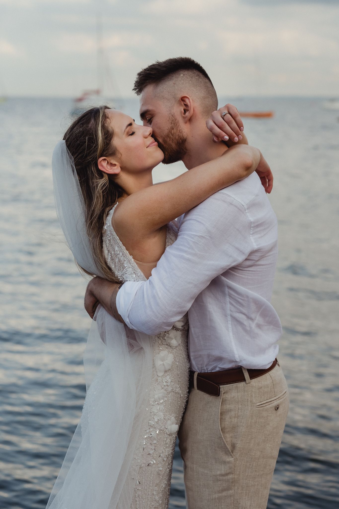 A romantic couple embraces and kisses by the ocean at sunset wearing wedding attire with a flowing veil.