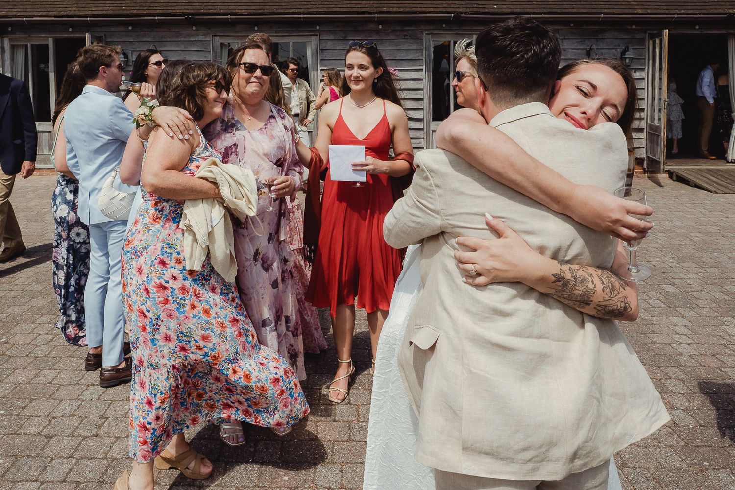 Wedding guests in floral dresses gather outdoors while two people share a warm embrace at a celebration.