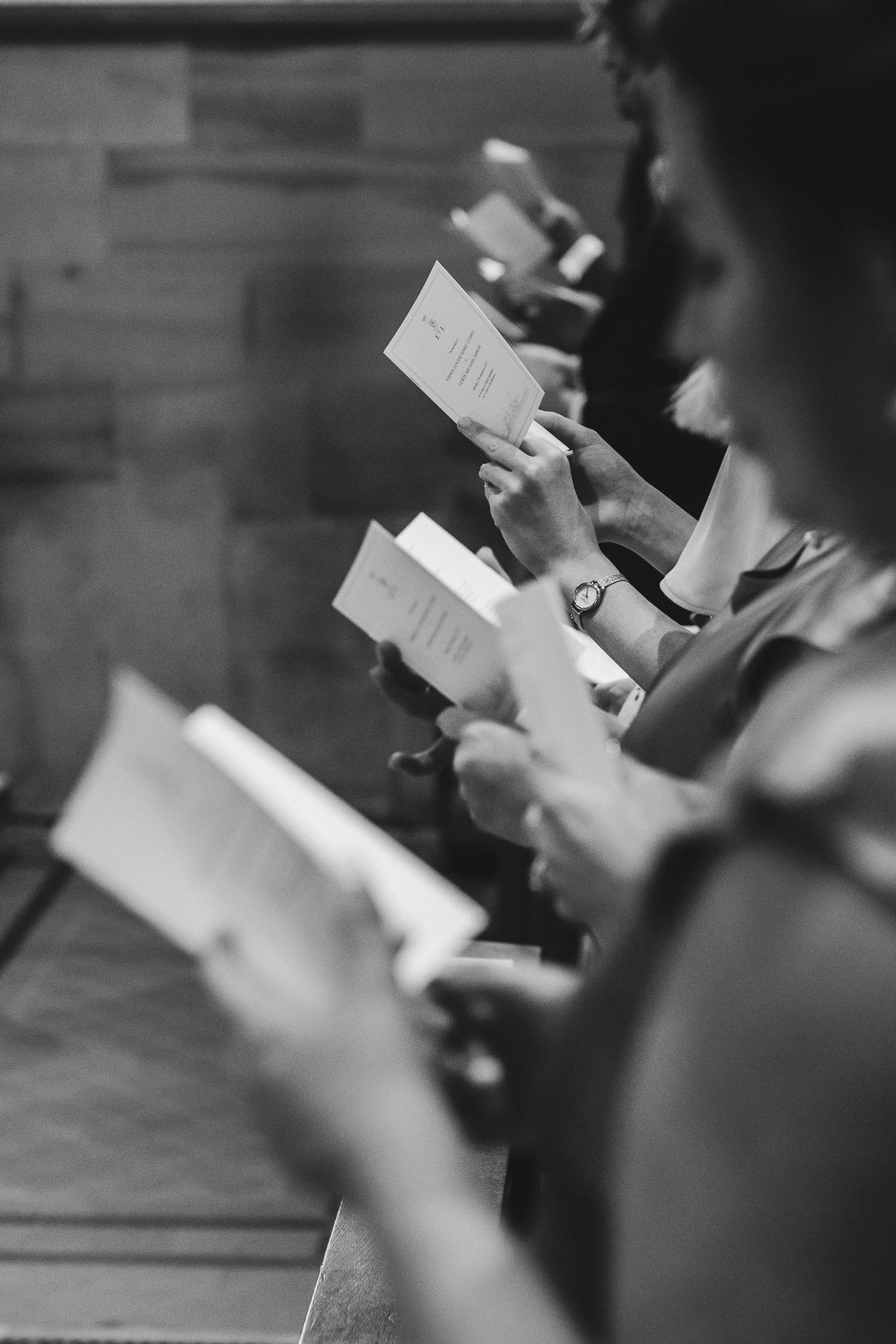 Multiple hands hold papers in a black and white scene showing a coordinated reading or performance.