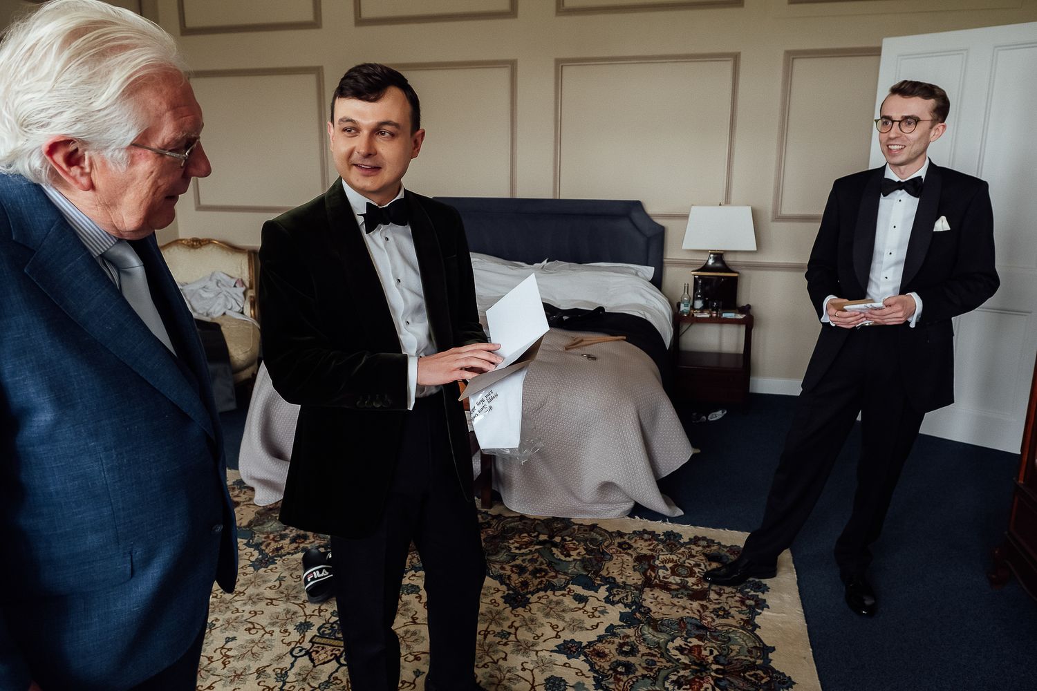 Three men in formal tuxedos preparing for a wedding ceremony in an elegant hotel room.