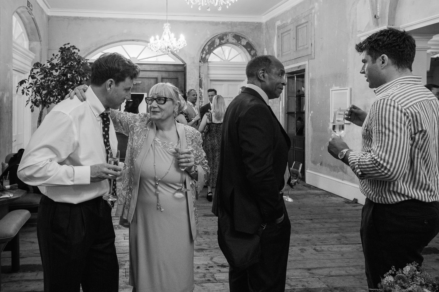 Guests laugh and chat while holding drinks at an elegant indoor wedding reception in a grand room with arched windows and a chandelier.