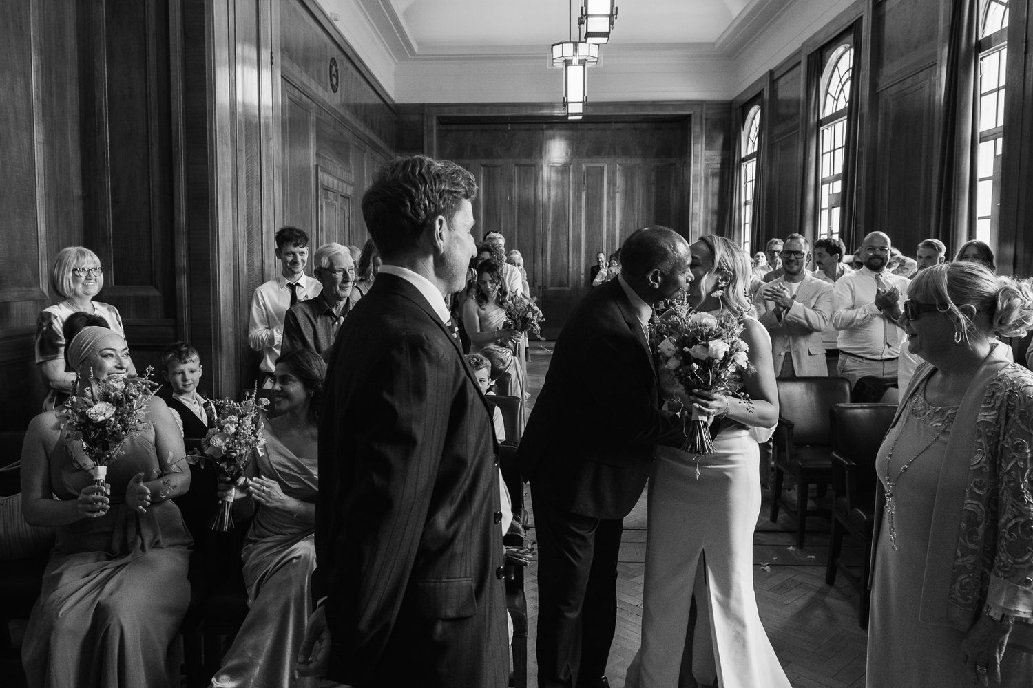 Black and white photo of a bride and groom sharing a kiss during a wedding ceremony as seated guests watch in a paneled room.