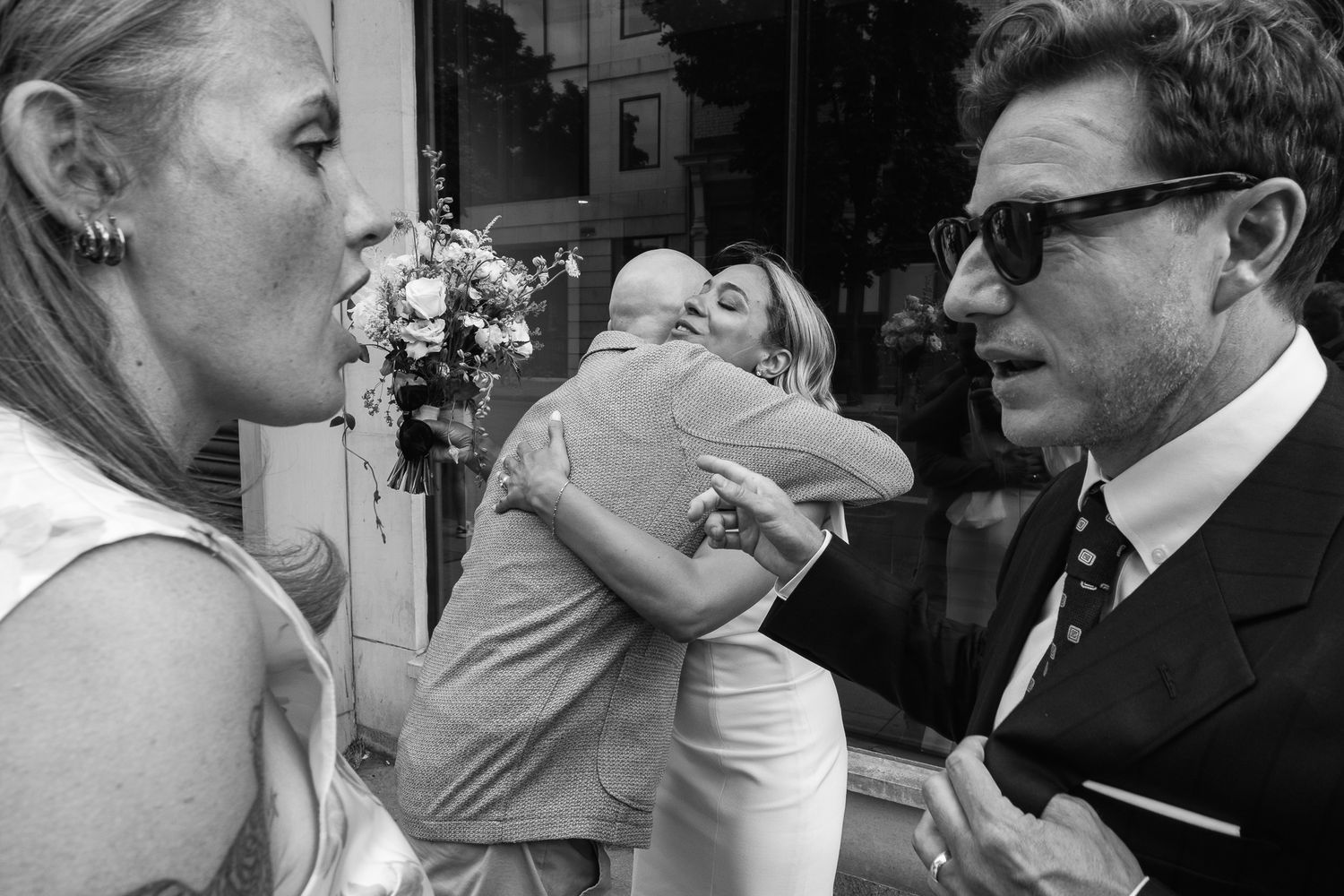 Black and white wedding photo of two guests embracing while a man in sunglasses and a tie and a woman in the foreground look on.