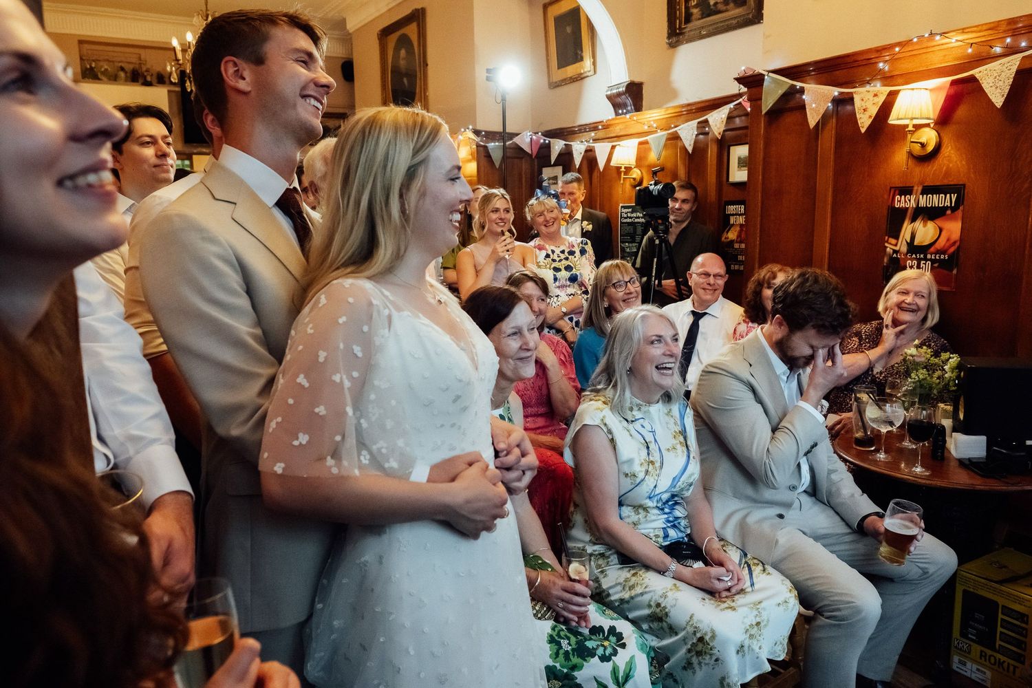 Smiling wedding guests in a cozy pub laugh and clap while a bride in a white dress stands beside a groom during a festive celebration.