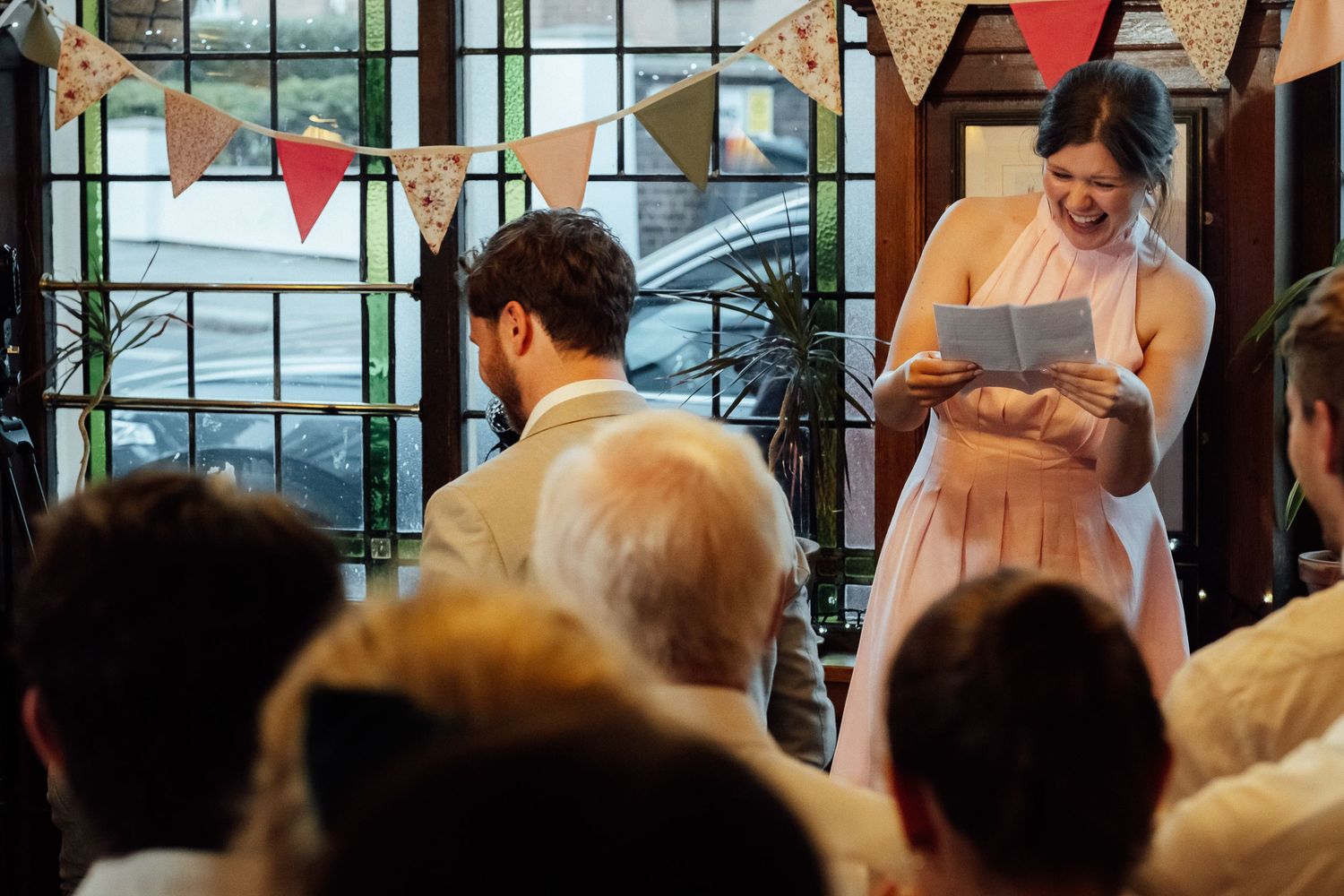 Woman in a pink dress laughs while reading from a paper to wedding guests under colorful bunting in a bright room.
