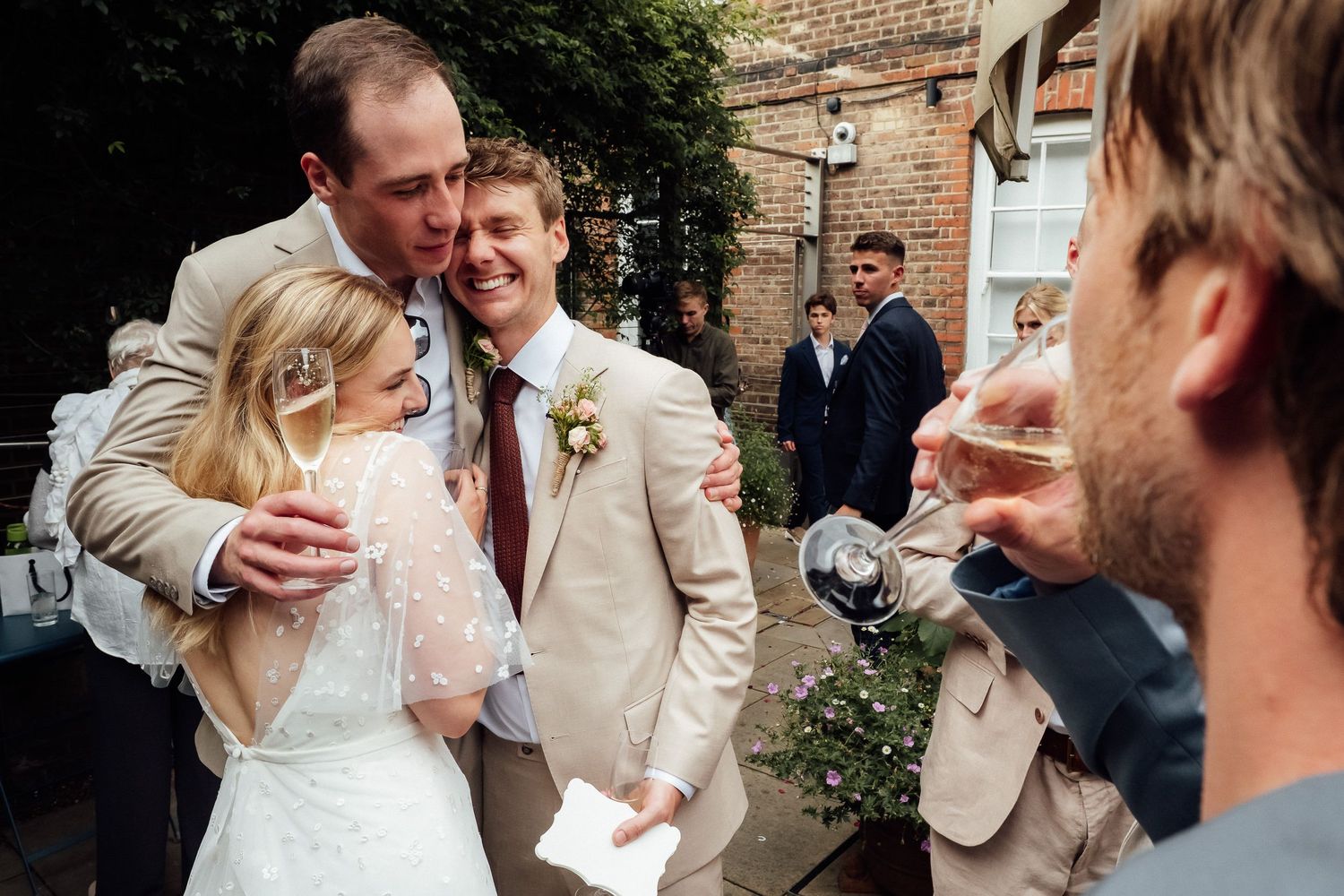 Smiling groom in a tan suit embraces a bride in a white lace dress while wedding guests celebrate with drinks in a courtyard.