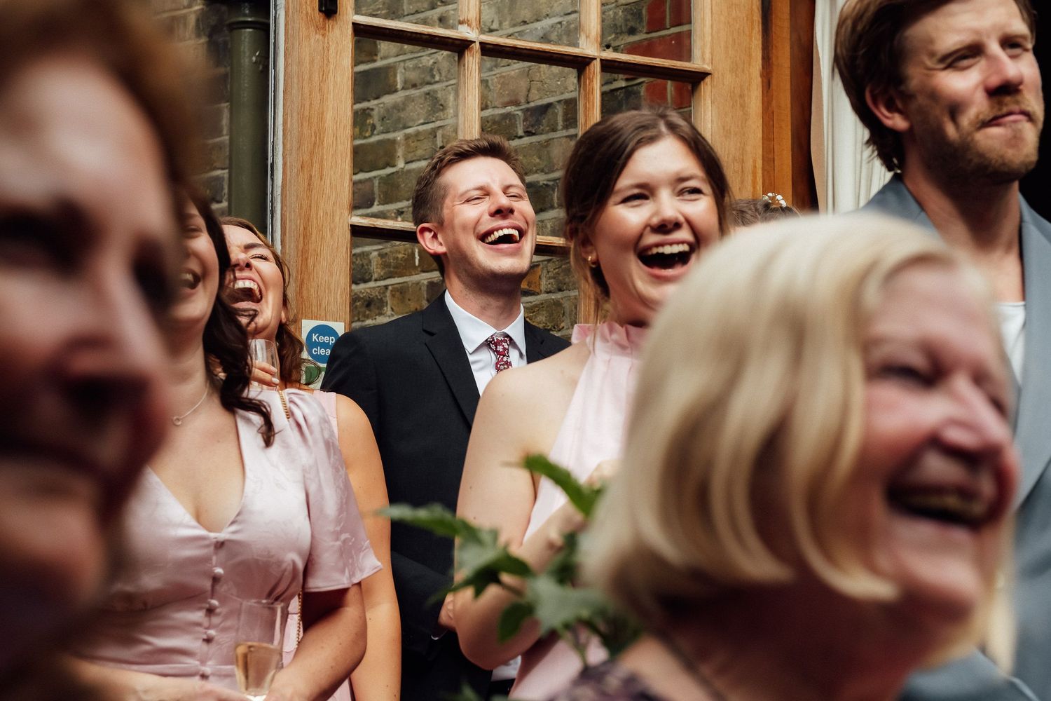 Group of smiling wedding guests in formal attire laughing together in front of a brick wall during a joyful celebration.