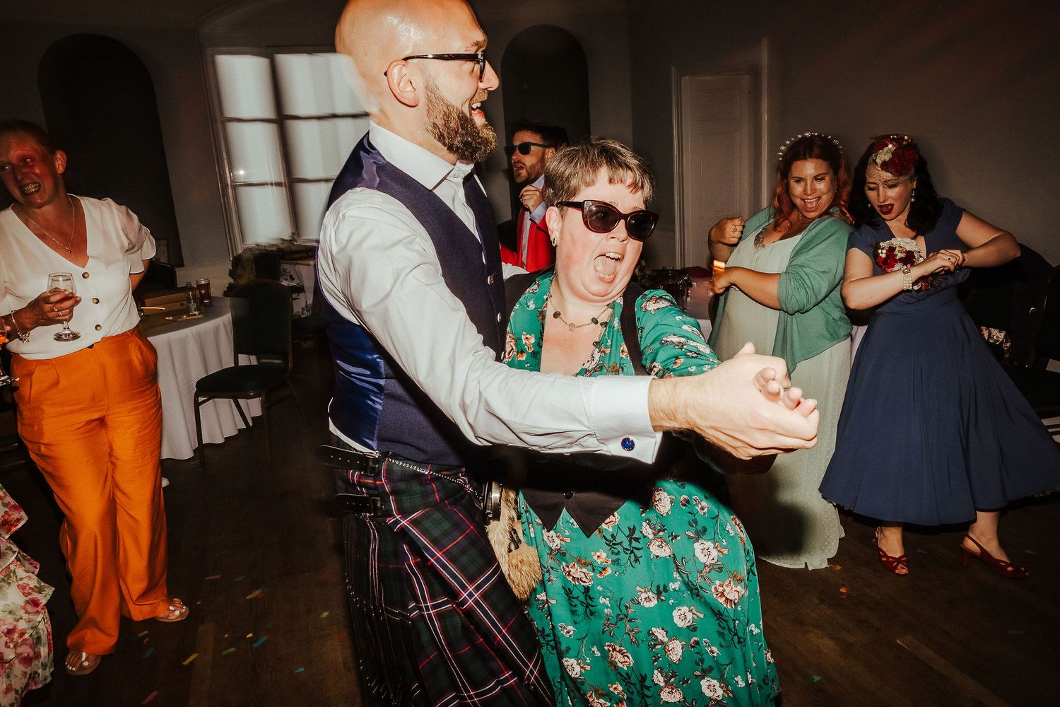 A bearded man in a kilt and waistcoat dances with a smiling woman in a green floral dress while guests watch at a wedding reception.