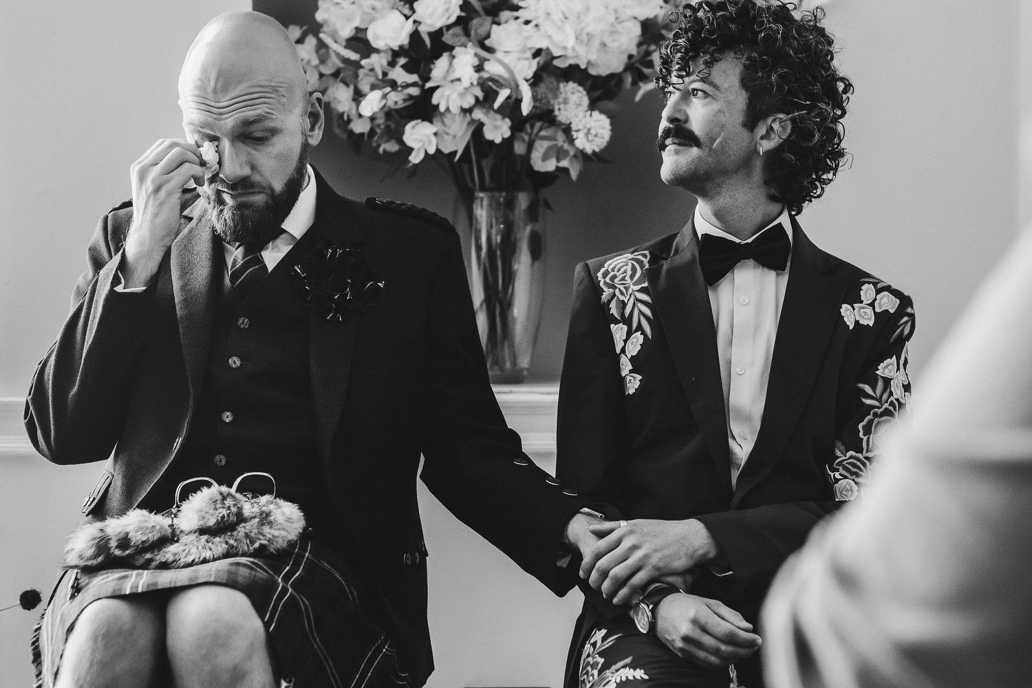 Two grooms in suits sit holding hands during a wedding ceremony as one wipes away a tear, with flowers in the background.