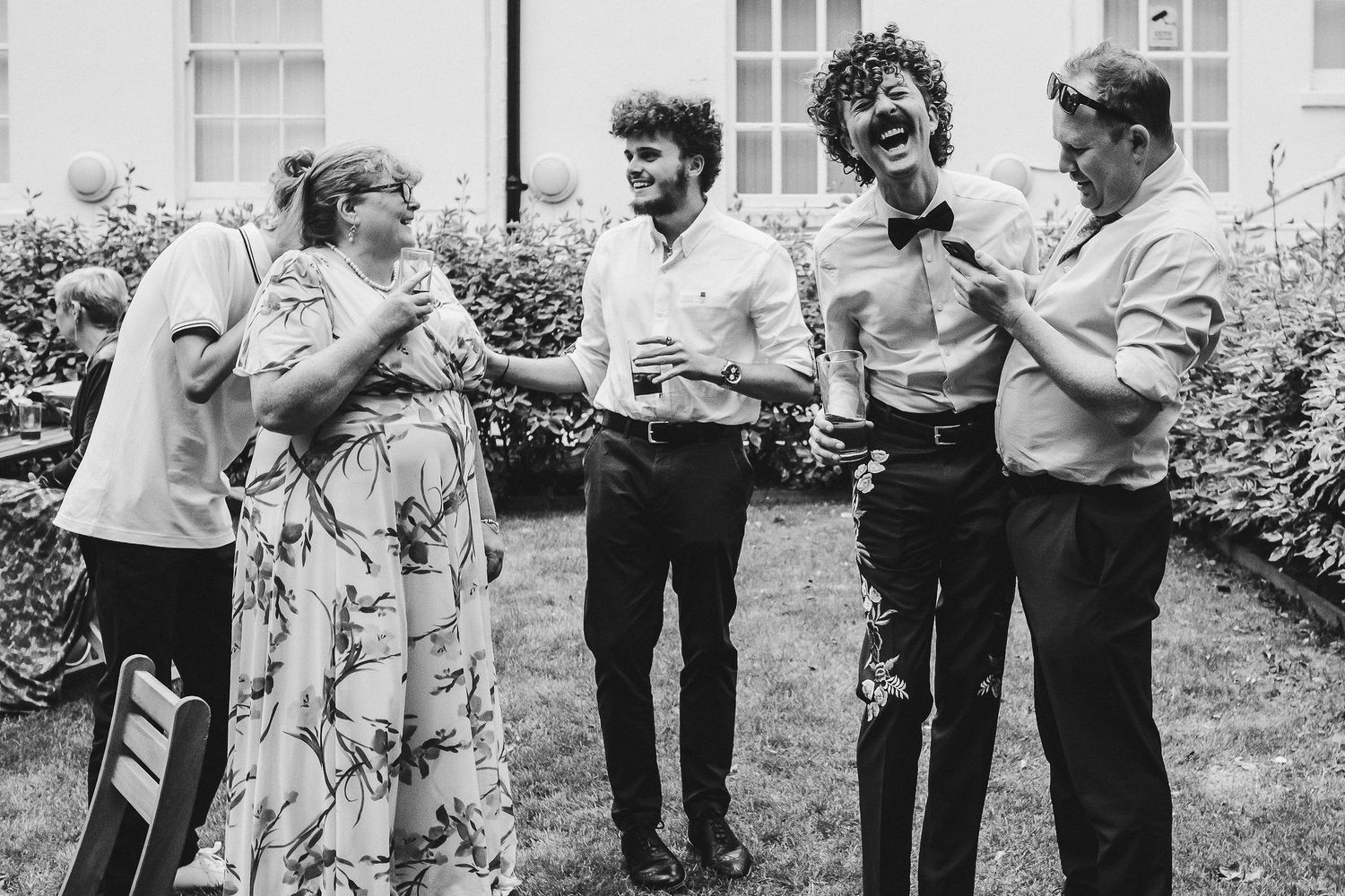 Black and white photo of guests laughing and chatting with drinks on a sunny garden lawn at an outdoor wedding celebration.