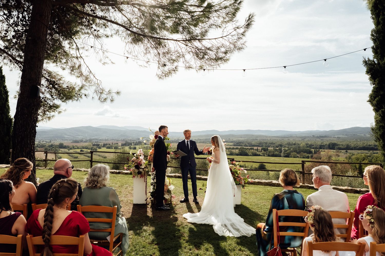 Bride and groom exchange vows during an outdoor countryside wedding ceremony as seated guests watch under a tree with rolling hills behind.
