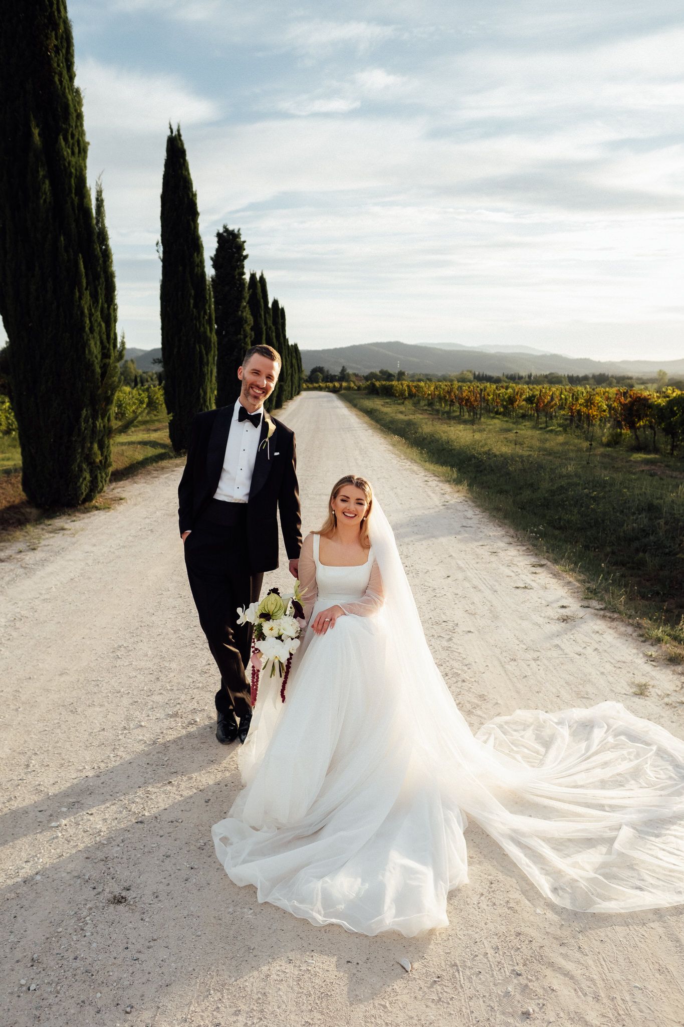 Bride in a flowing white gown and veil holds a bouquet beside a groom in a tuxedo on a gravel road lined with cypress trees and vineyards.