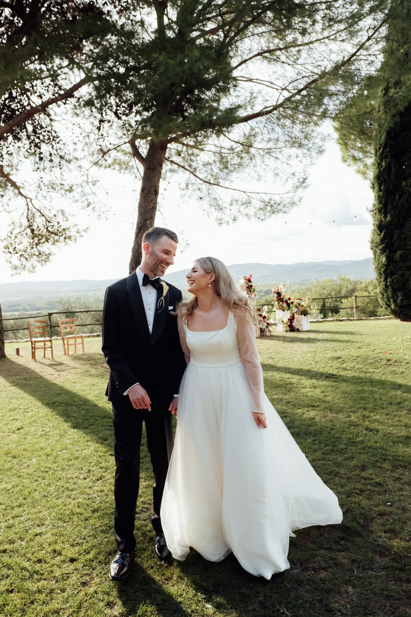 Smiling bride in a white gown and groom in a black tuxedo hold hands on a sunny lawn with pine trees and rolling hills behind them.