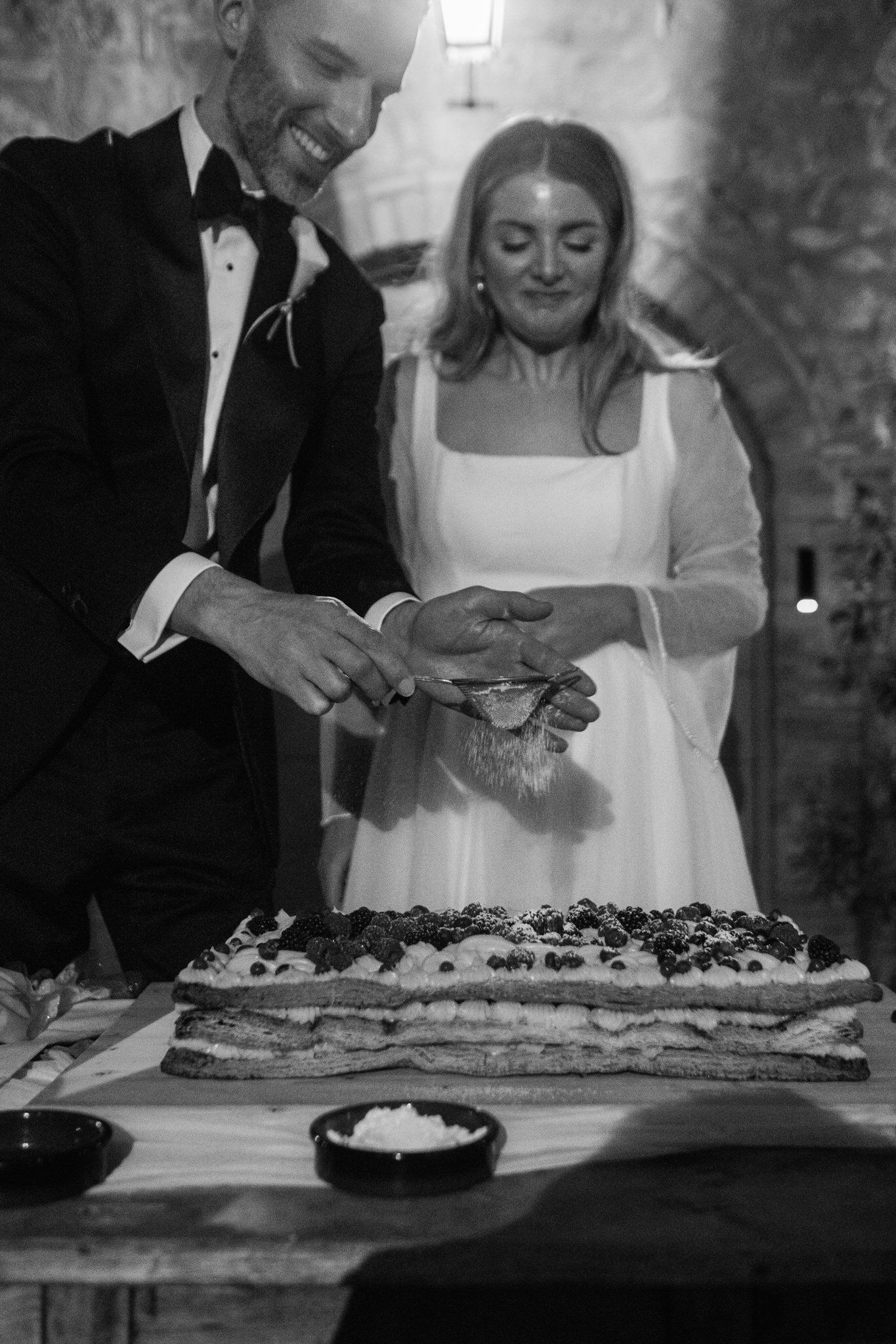Smiling bride and groom in formal wedding attire sprinkle sugar over a layered berry wedding cake in a rustic stone venue.