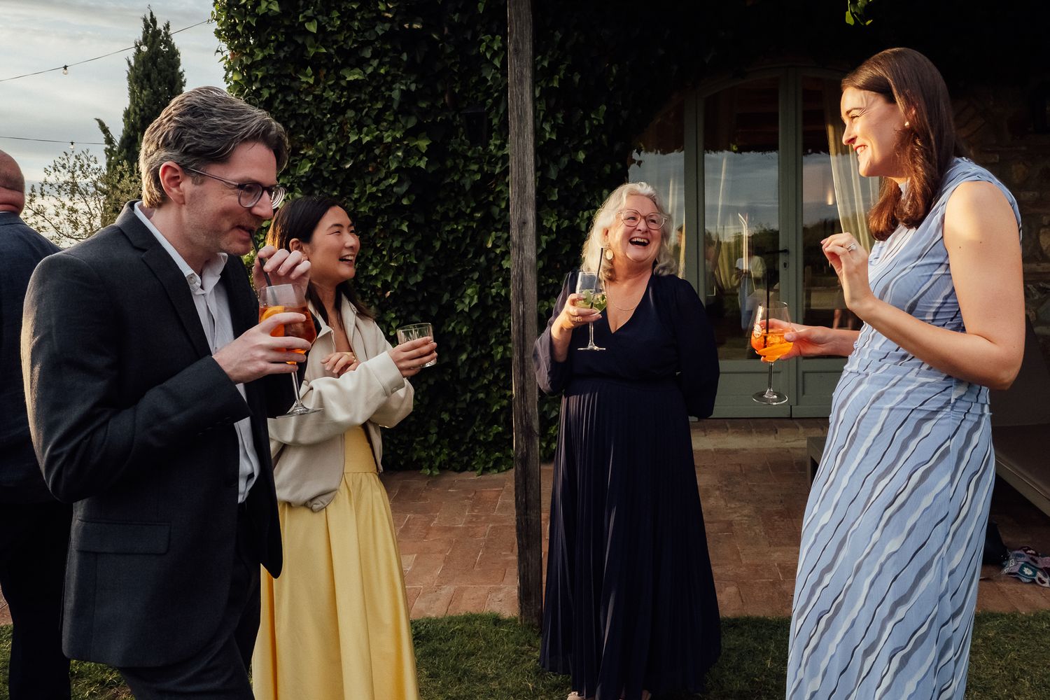 Four guests laugh and chat while holding drinks at an outdoor evening gathering beside an ivy-covered building.