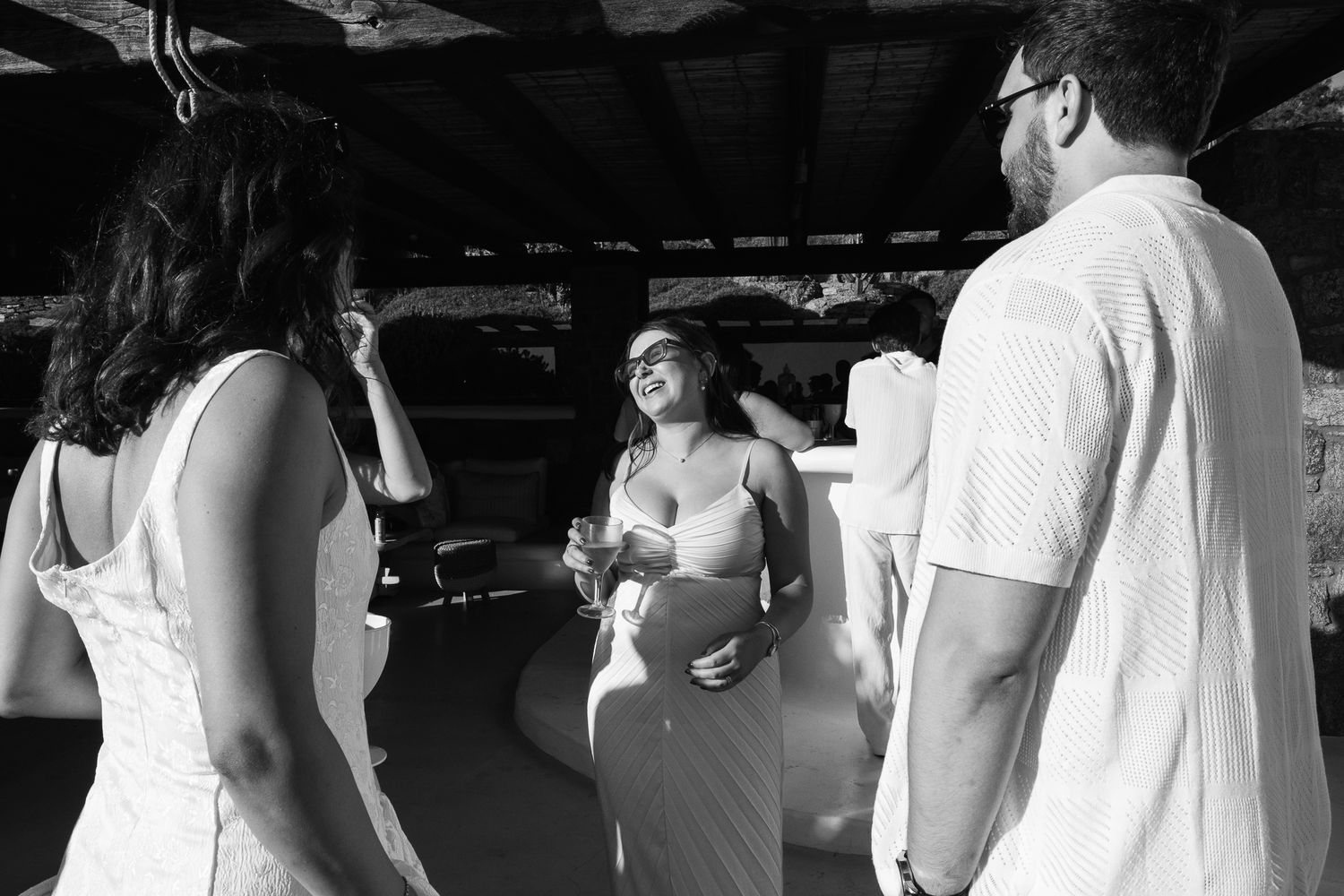 Black and white photo of three people conversing at an outdoor wedding reception, woman in center smiling holding a drink.