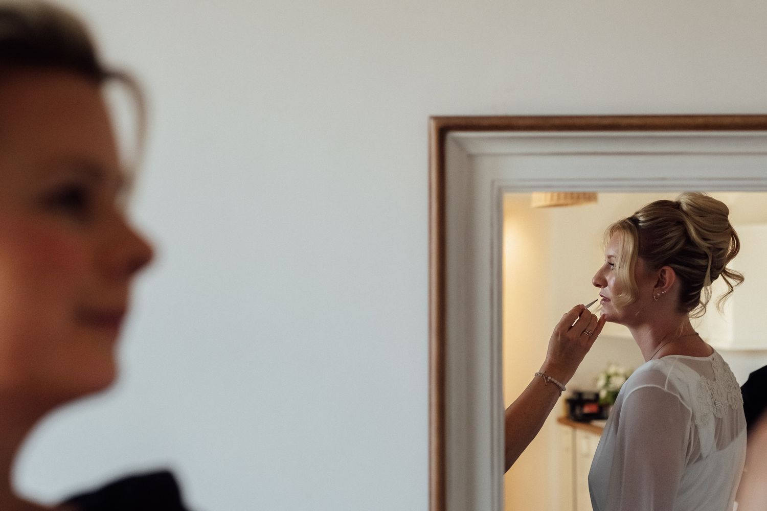 Bride getting makeup applied by artist, reflected in mirror, with blurred bridesmaid in foreground at wedding preparation.