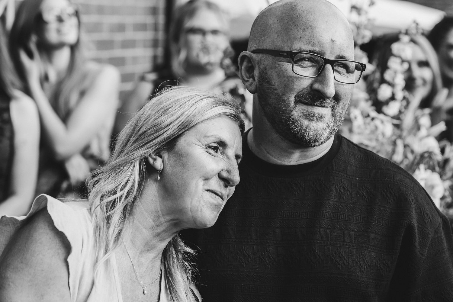 Black and white candid portrait of a couple sharing an intimate moment outdoors with others visible in background.