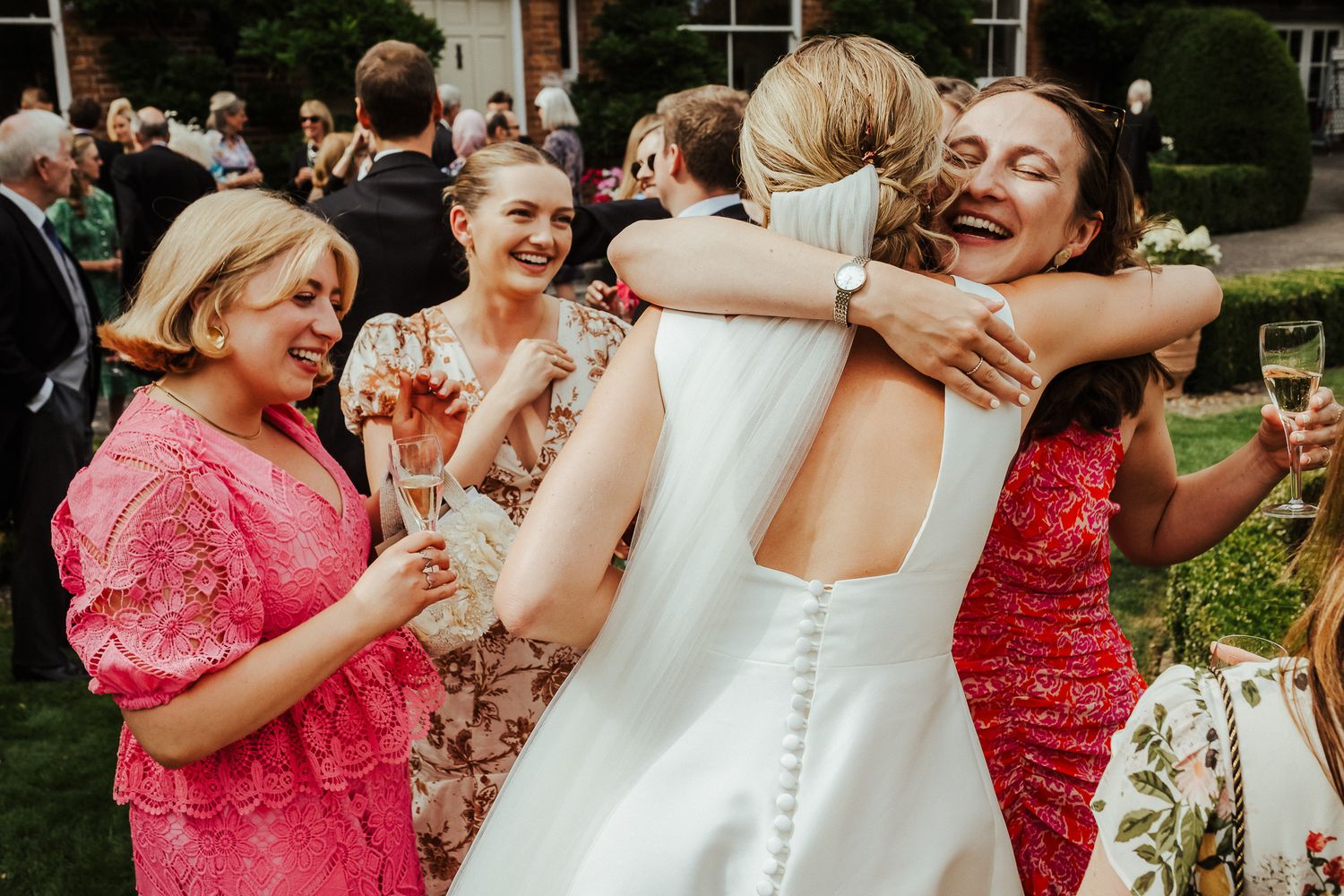 Guests celebrate at an outdoor wedding reception with laughter and hugs among pink floral decorations.