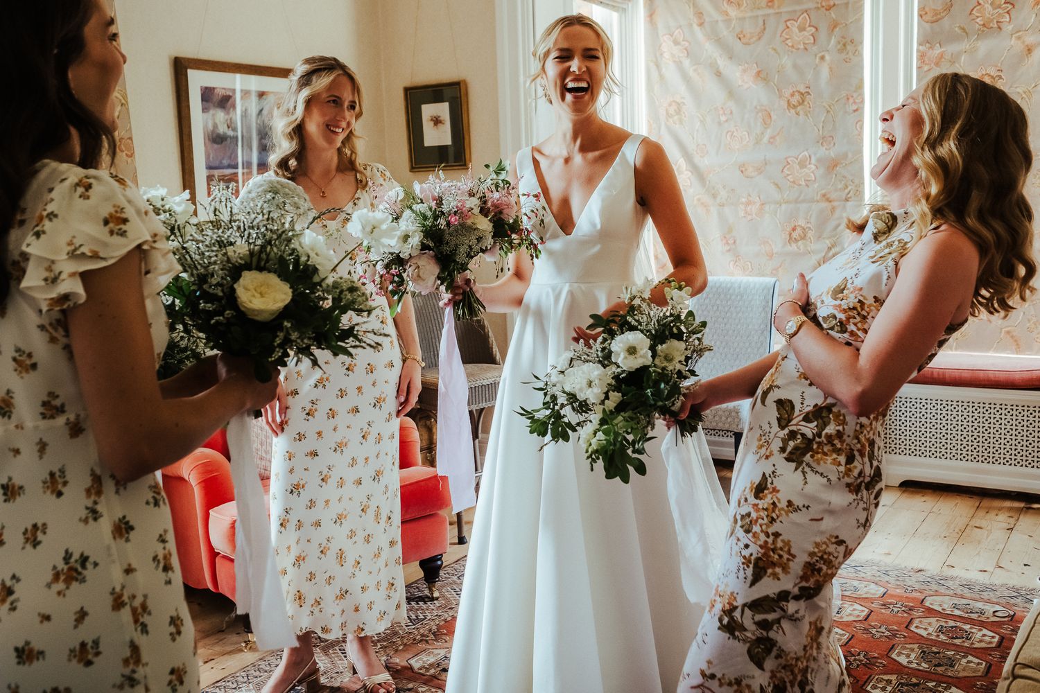 A bride in a sleeveless white gown shares a laugh with her bridesmaids in floral dresses.