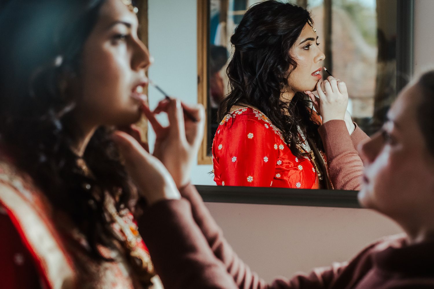 A woman in a red polka dot dress applies makeup while looking in a mirror during traditional preparations.